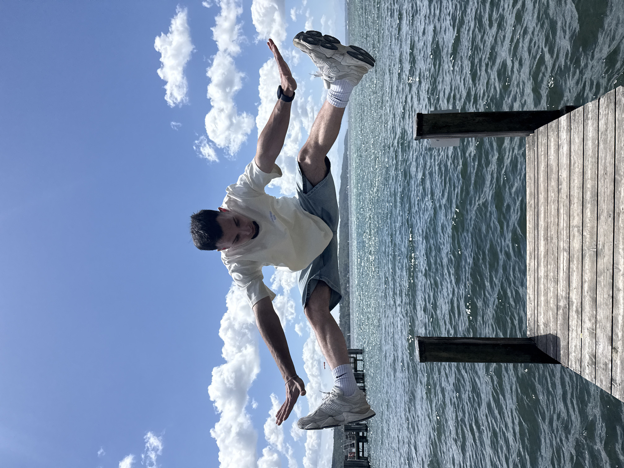 A man is captured mid-air in a wide split jump, suspended above a wooden dock. He wears a light-colored t-shirt, denim shorts, white socks, and athletic shoes. His arms are outstretched, and his head is tilted downwards, looking towards the water. The background features the calm, blue waters of the Starnberger See, with a distant shoreline of trees and buildings under a bright blue sky dotted with fluffy white clouds. A pier with wooden pilings extends into the lake. The scene conveys a sense of freedom and athleticism against a scenic natural backdrop.