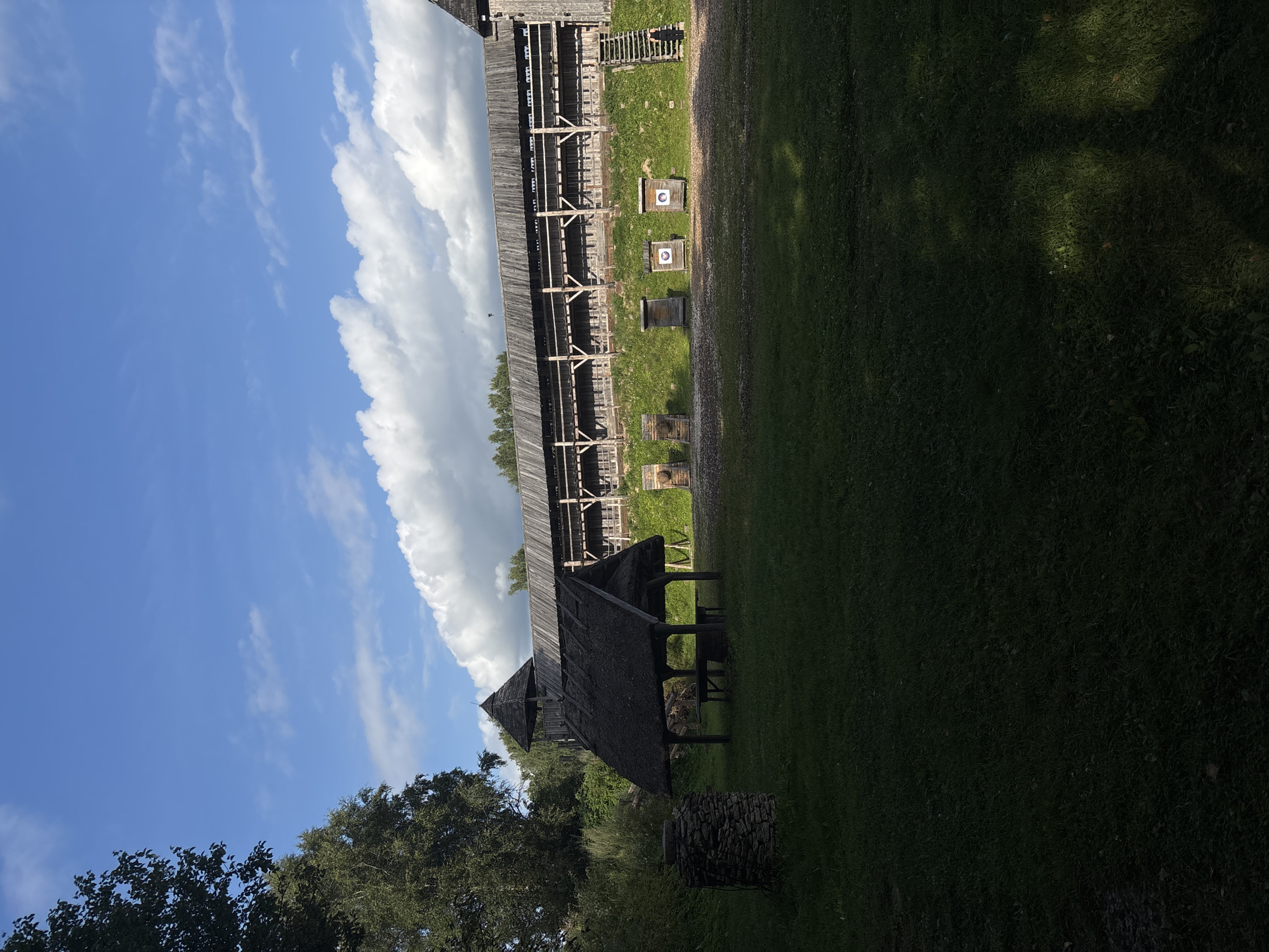 The image depicts an outdoor scene at a historical reenactment site, likely a fortress or palisade. In the foreground, a large expanse of grass is partially shadowed by trees on the left and a structure on the right. To the left, a small, dark-colored stone structure stands in the grass. Center-left, a rustic open-sided shelter with a thatched roof is visible, containing wooden benches and a table. Behind the shelter and extending across the middle ground is a tall, wooden palisade wall, characteristic of historical fortifications, with a wooden watchtower or turret visible to the left of it. In front of the palisade, a grassy slope leads up to the wooden structure. Several archery targets, some with circular bullseyes, are set up on wooden stands on the slope. To the far right, a wooden staircase leads up to the palisade. A lone person, dressed in black, is ascending these stairs. The sky above is a vibrant blue with scattered white, fluffy clouds. The overall atmosphere is one of a sunny day in a historical outdoor setting. The location is identified as the historical reenactment grounds in Saula, Estonia.