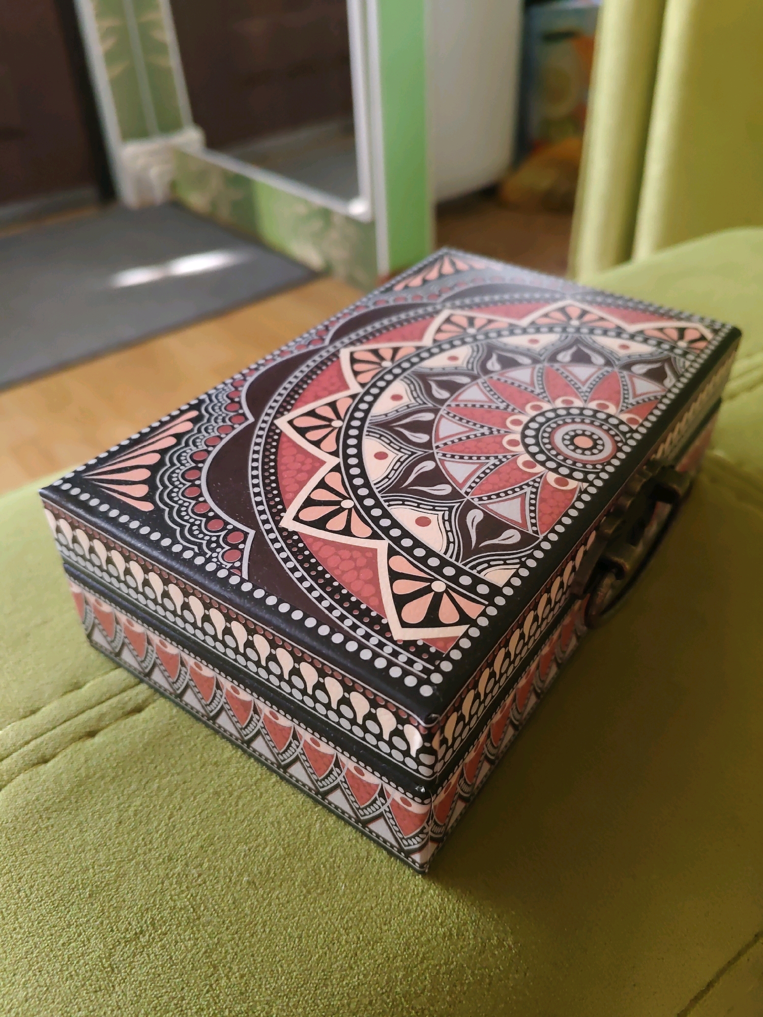 The image shows a decorative rectangular box with intricate mandala-like patterns in shades of brown, pink, and white, resting on a textured, lime-green surface. The box has a metallic clasp on its right side. The background is softly blurred, revealing what appears to be an indoor setting with hints of wooden flooring, a doorway, and a green curtain or fabric. There are no people or animals visible in the image, and no specific elements indicate a location within the city of Rostov-on-Don, Russia. The lighting suggests an indoor environment, possibly with natural light entering from a window.