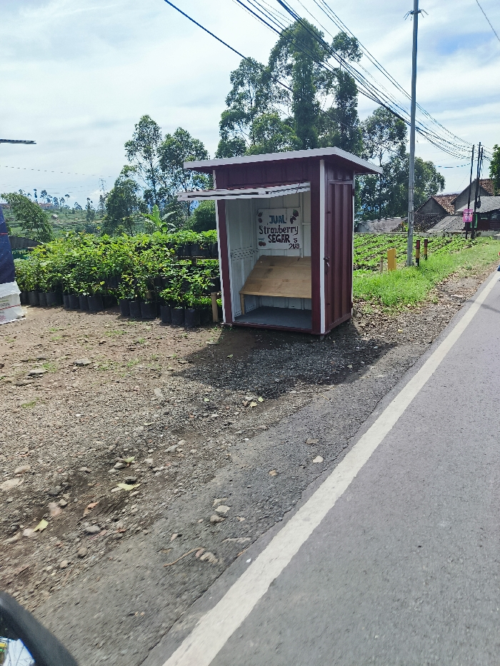 The image shows a small, red and white, wooden shack-like structure on the side of a paved road. The shack is labeled with the words “JUAL Strawberry SEGAR” written in black lettering. There is a small, wooden shelf in the shack, along with a small wooden bench. The road is bordered by a white line and a dirt shoulder. Behind the shack is a row of small, green bushes and trees. The image appears to be taken in a rural area, as there are no other buildings or signs of human activity besides the shack. The coordinates provided, -7.122 latitude and 107.429 longitude, place the image near Rancabali, Indonesia. The scene is captured from a low angle and appears to have been taken in the daytime. The sky is clear and there are no clouds. The image is in focus and well-lit.