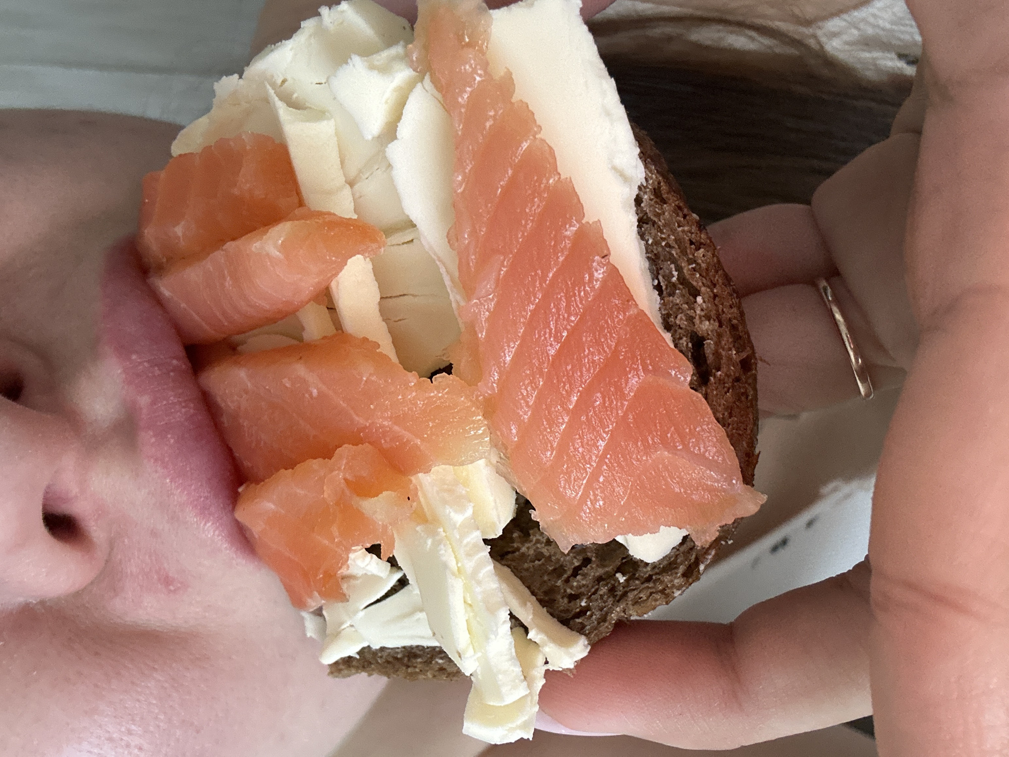 A close-up image shows a person holding a piece of dark bread topped with cream cheese and slices of salmon. The person is bringing the bread close to their mouth. The location is not identifiable in the image, nor is there any indication it is in Chaikovsky, Russia. The person is wearing a ring on their finger. The sandwich appears to be held up to the person's mouth.