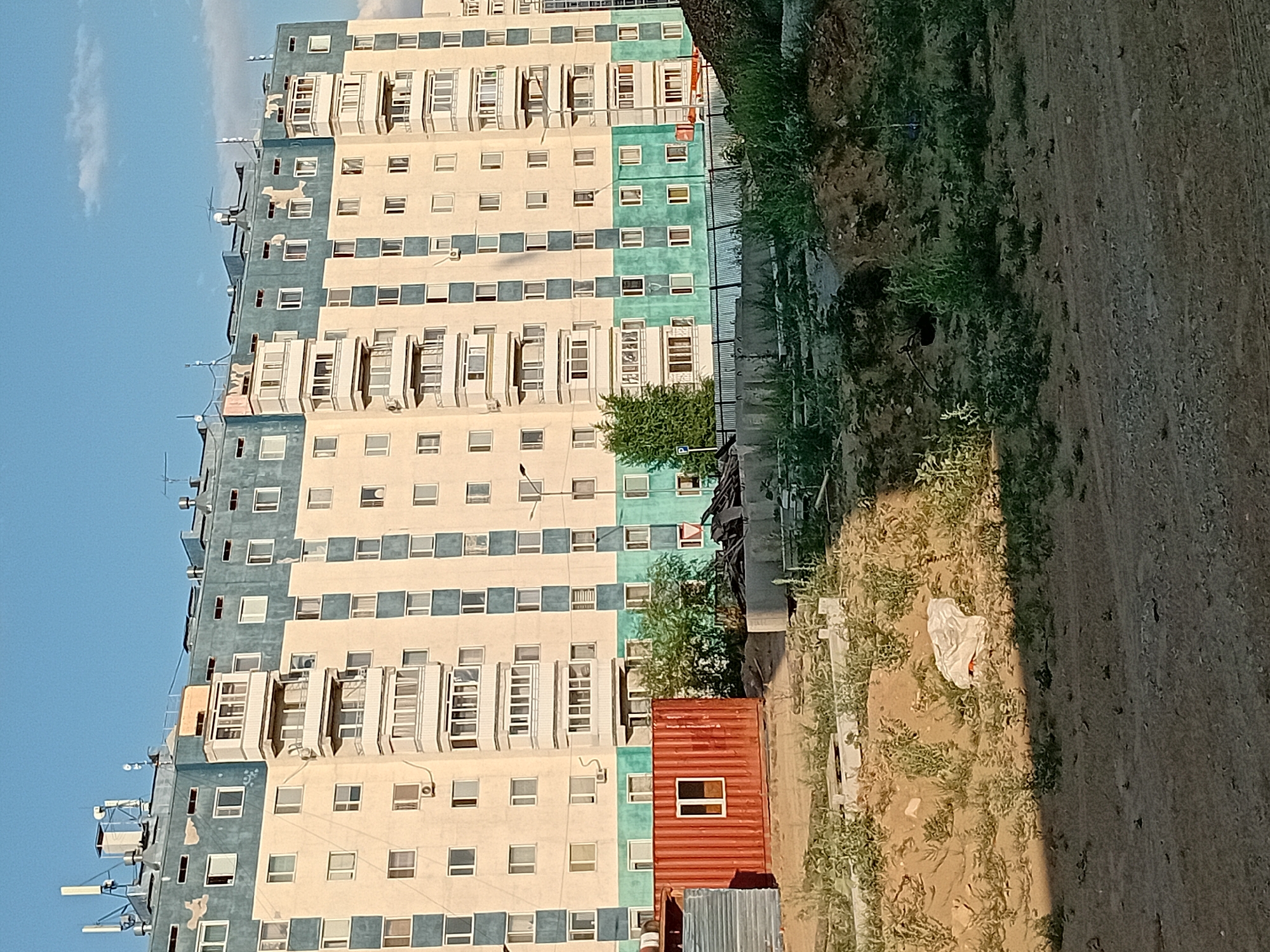 An outdoor, eye-level, medium shot shows a multi-story apartment building in the city of Semey, Kazakhstan. The building is tall and narrow, with a cream-colored facade and a teal-colored top section. Many windows and balconies are visible on the building. In the foreground, there is a construction site with piles of dirt, some debris, and a red shipping container with a white-framed window. A chain-link fence runs along the middle ground, separating the construction site from the building. The sky above is a clear blue with a few wispy white clouds. The lighting suggests it is late afternoon.