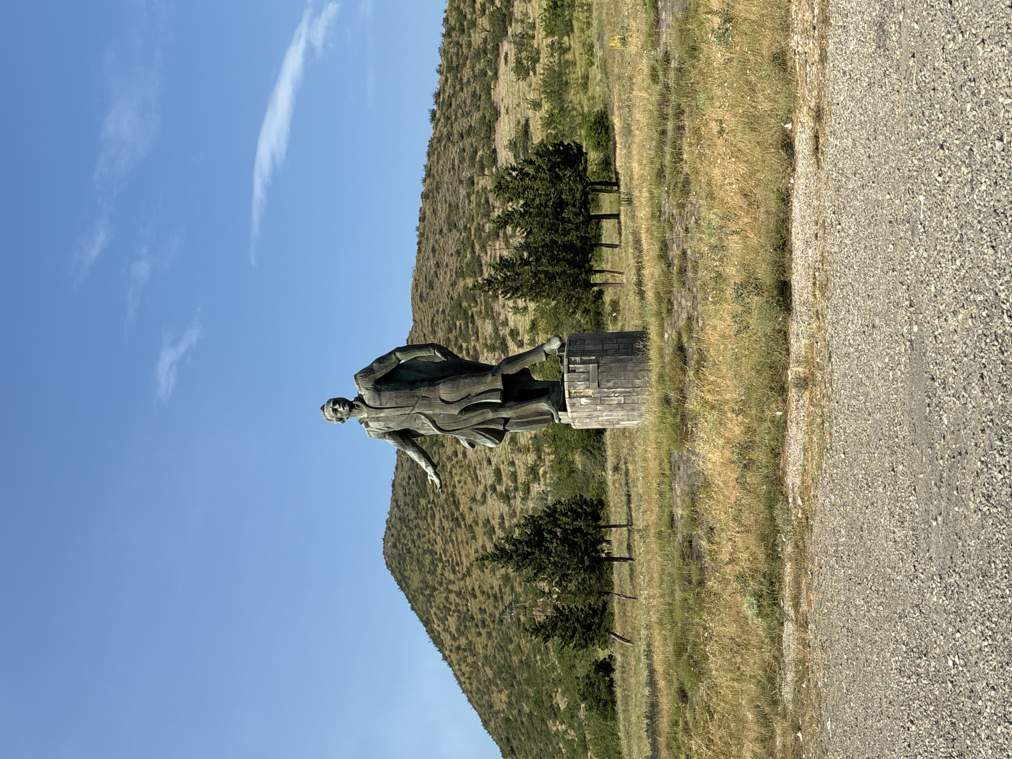 In Mukhatgverdi, Georgia, a large bronze statue of a man in a suit stands prominently on a concrete pedestal in a grassy field. The statue depicts the man with his left arm extended forward and his right arm bent at the elbow. Behind the statue, a barren, rolling hill is covered in sparse vegetation. Several evergreen trees are scattered in the foreground and midground. A dirt road is visible in the lower portion of the image. The sky is clear blue with a few wispy clouds.