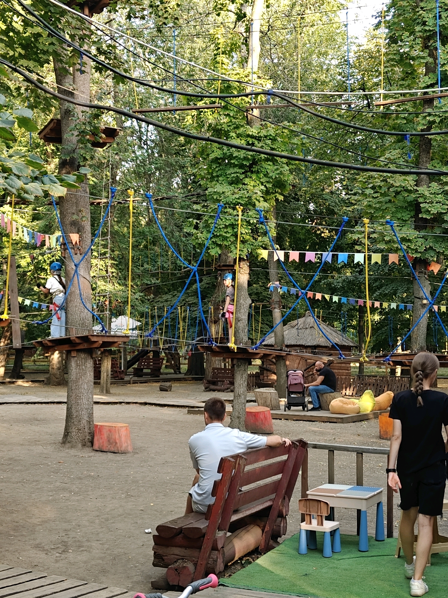 This image depicts an outdoor adventure park with rope courses suspended between trees. In the foreground, a man sits on a wooden bench, facing away from the camera. To the right, a woman with braided hair walks away from the camera. A child-sized table and chair set is visible on a green mat. In the midground, a rope course is in operation, with two children in safety gear navigating the obstacles. A thatched-roof structure and other park amenities are present. The background is dominated by lush green trees, suggesting a park or forest setting. The scene is likely located in the city of Rostov-on-Don, Russia, given the information provided.