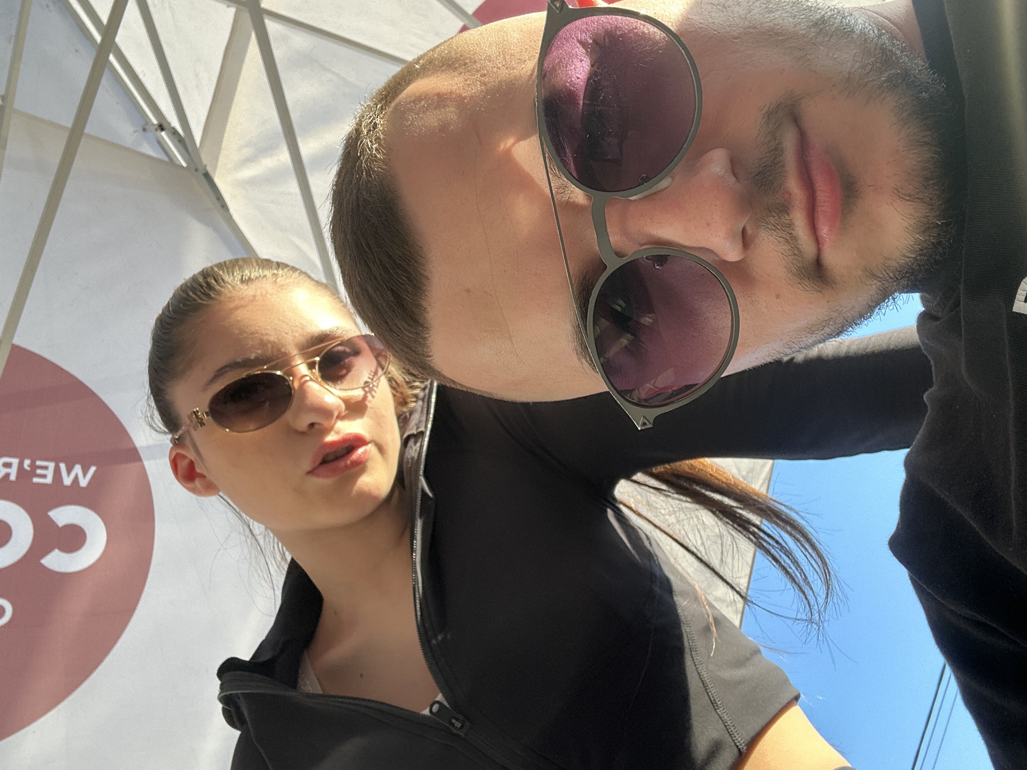 An eye-level selfie captures two individuals from a low angle on a bright, sunny day in Plovdiv, Bulgaria. On the left side of the frame, a woman with dark hair pulled back, wearing a black zip-up top and ornate golden sunglasses, gazes forward with a slightly open mouth. Her face is well-lit, showing prominent cheekbones and full lips.
Dominating the upper and right portions of the frame is a man, whose head is tilted and inverted relative to the woman, facing the camera. He wears dark, round sunglasses and has short dark hair with a beard or heavy stubble. His dark hooded garment blends with the shadows.
The background features a white outdoor umbrella or canopy structure, its support beams clearly visible against the bright sky, suggesting an outdoor setting like a cafe or market. Behind the woman, a red circular sign with white, mirrored text is visible. The legible parts of the text read "М.Е.В." (appearing as "В.Е.М." due to mirroring) and "СО" (appearing as "ОС" or "ƆО"). A clear blue sky is visible in the lower right and upper right corners, indicating fair weather. The strong sunlight casts highlights and shadows on the subjects, emphasizing their features and sunglasses.