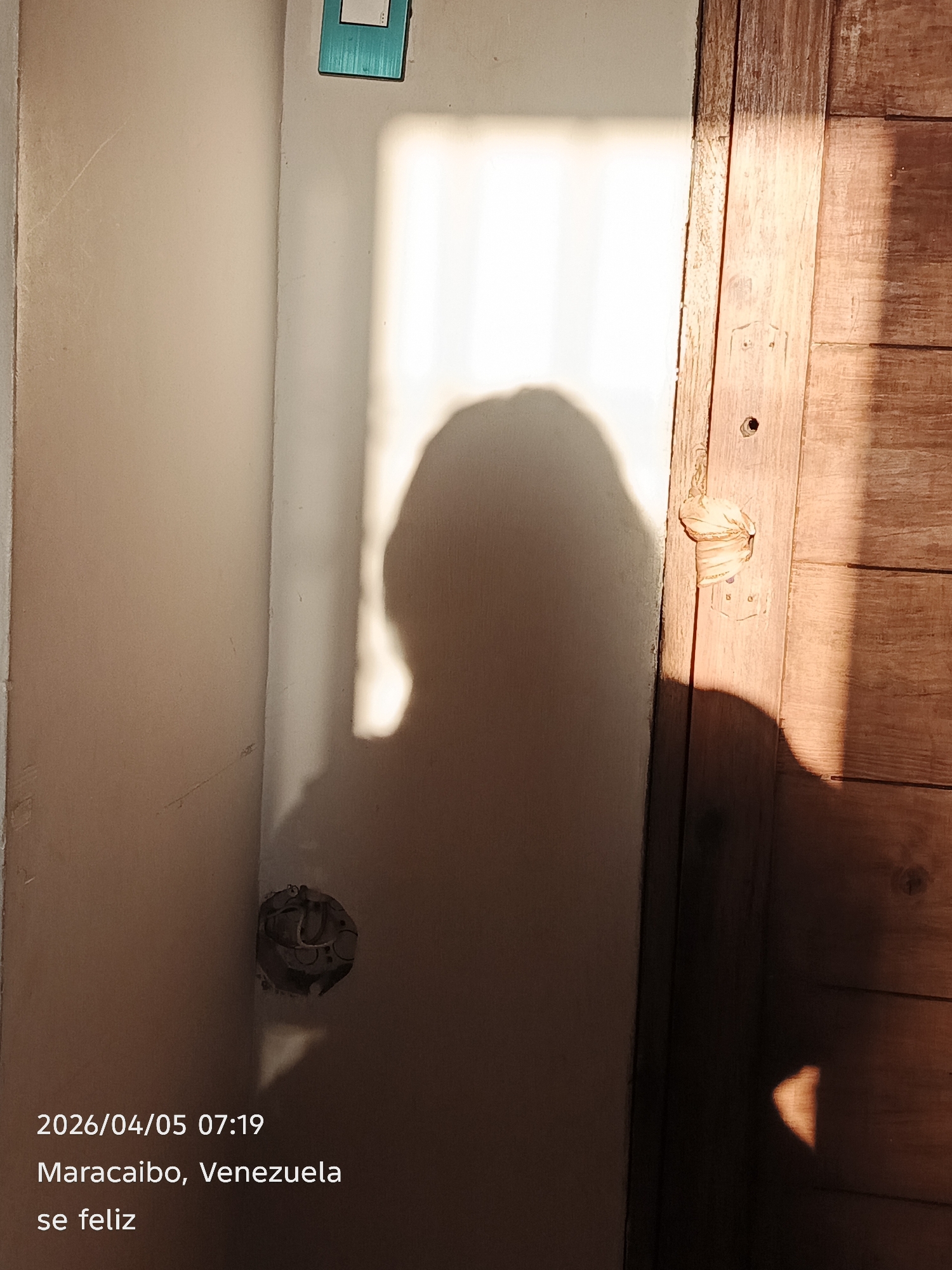 The image shows a shadow of a person on a wall next to a wooden door. The wall is painted a light color, and there's an electrical outlet on the lower left side of the wall. The wooden door is on the right side of the image. The image also includes text at the bottom indicating the date and time (2026/04/05 07:19), the location (Maracaibo, Venezuela), and the phrase "se feliz" (be happy).