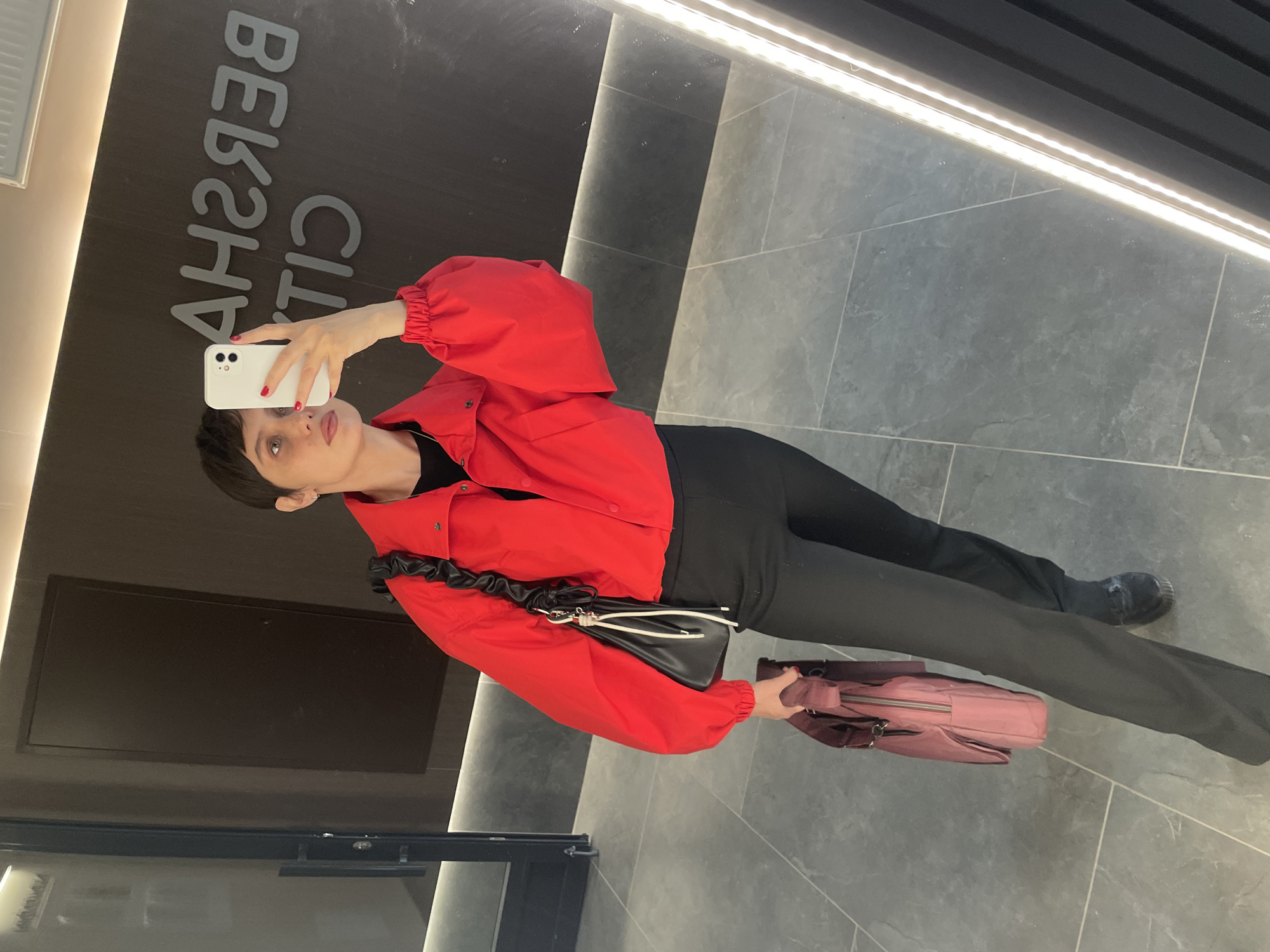 A woman in a red jacket and black pants is taking a selfie in front of a mirror. The mirror reflects the interior of what appears to be a retail space, with the word "BERSHKA" visible in the reflection. The woman is holding a white smartphone in her right hand and a pink bag in her left hand. She is also carrying a black purse with a ruffle strap. The floor is tiled in grey. The location is Izhevsk, Russia.