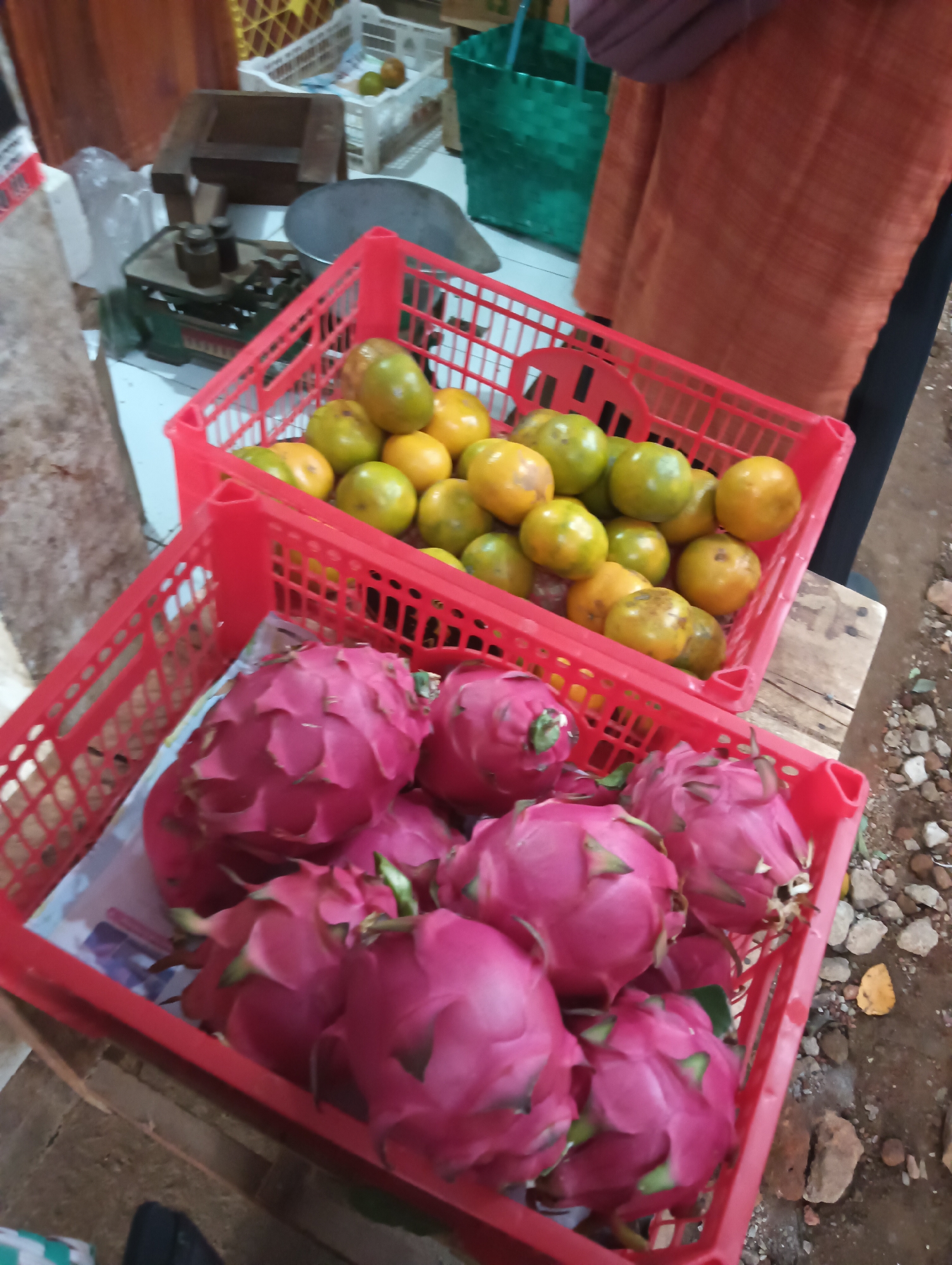 A street market stall in Cluwak, Indonesia, displays two red plastic crates filled with fruit. The front crate contains several vibrant pink dragon fruits. Behind it, a second red crate is packed with a mix of green and yellow oranges. In the background, a vintage scale with weights sits on a surface, alongside other market items that are out of focus. A person wearing an orange garment is partially visible in the upper right corner of the image. The ground appears to be unpaved.