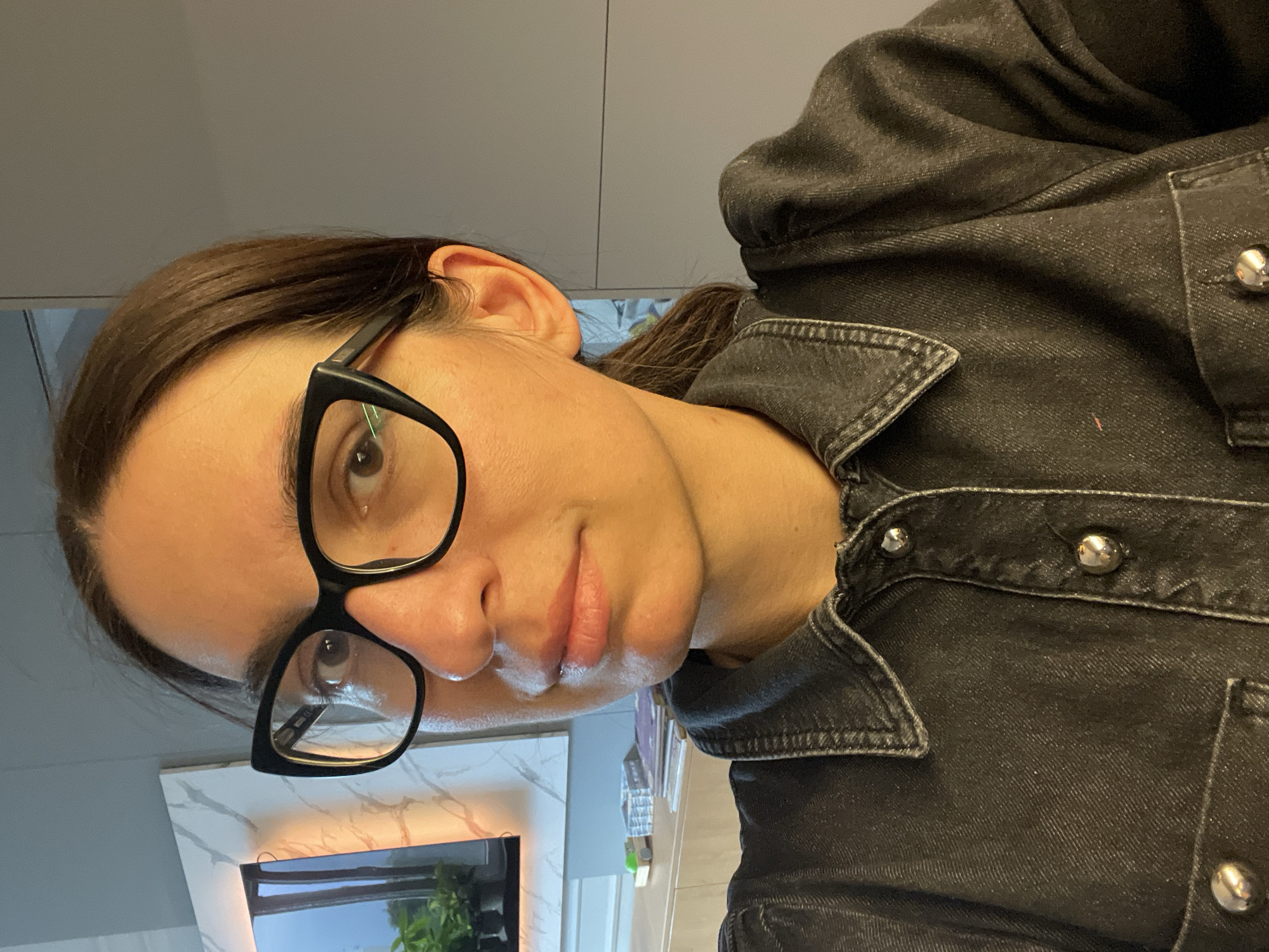 The image is a close-up, selfie-style portrait of a person, positioned with their head tilted. The subject, likely female, has dark brown hair pulled back, and is wearing black, thick-rimmed glasses. Their brown eyes look directly towards the camera, conveying a neutral to slightly contemplative expression with a hint of a smile.
The person is dressed in a dark, possibly black or dark gray, denim-like shirt or jacket. Notable details of the garment include silver metallic snap buttons on the collar and front placket, along with visible stitching that gives it a rugged, casual aesthetic.
The background indicates an indoor setting, likely a modern kitchen or living area. A light gray wall or cabinet is visible behind the subject's head. Below, there's a white and gray marbled surface, possibly a countertop or decorative panel, featuring a warm-toned linear light strip. Further down, a dark reflective surface shows a blurred reflection of green plants or foliage, suggesting a window or display screen is in the vicinity.
The activity taking place is a direct portrait or selfie. The lighting is soft and even, suggesting an interior setting with adequate illumination, possibly from natural light or internal fixtures. No specific time of day or weather conditions can be definitively determined from the indoor scene. The subject appears calm and composed. No visible text is present in the image. The provided location context is Sofia, Bulgaria.