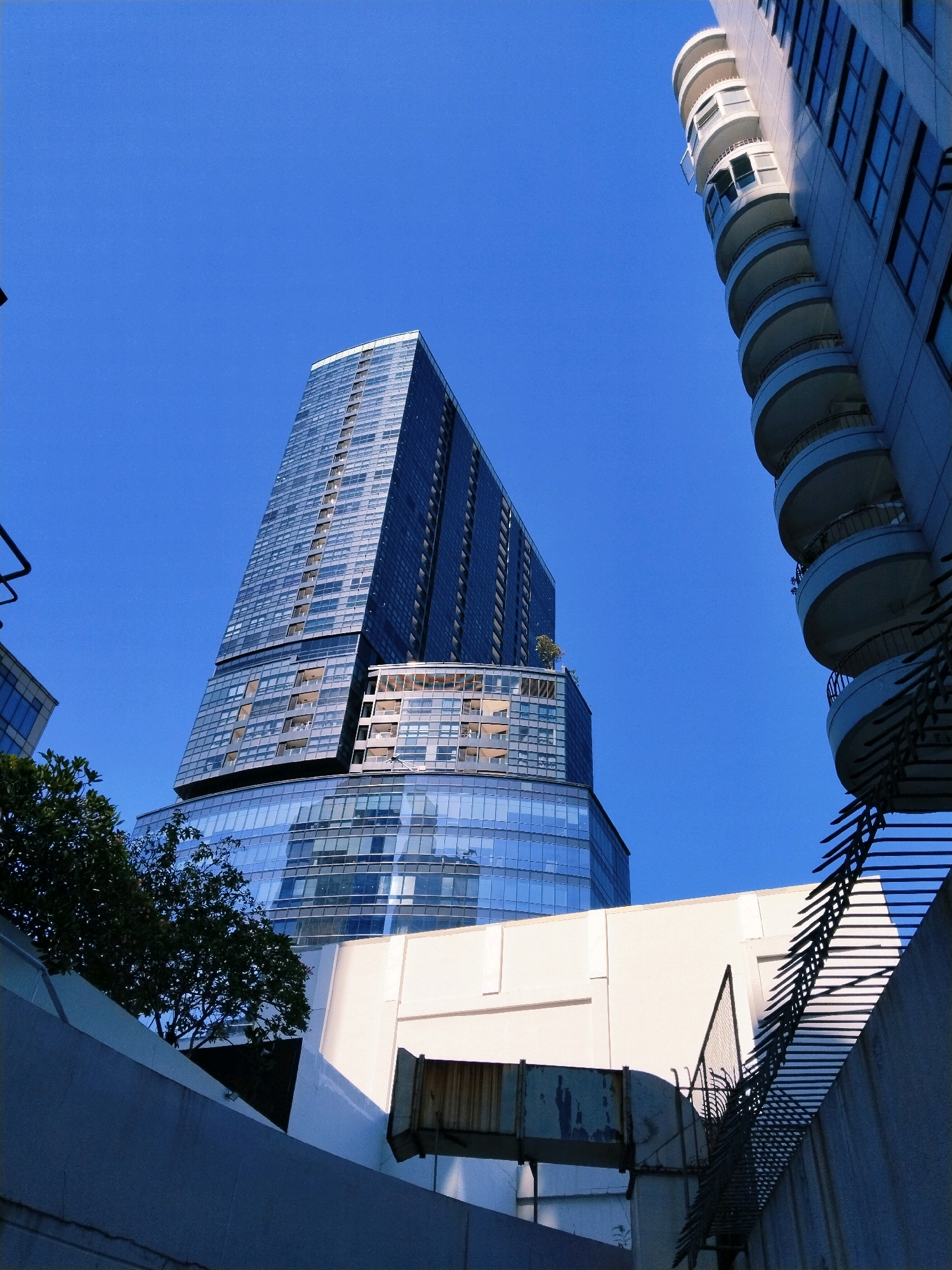 The image, taken from a low angle in Surabaya, Indonesia, showcases a clear blue sky above a cluster of modern buildings. Dominating the frame is a tall, sleek skyscraper with a glass facade, reflecting the surrounding environment. To its right, another building features a series of curved balconies, adding a distinct architectural element. In the foreground, a light-colored wall with a grated structure casts a shadow, hinting at an urban setting. Trees with dark green foliage are visible to the left, providing a natural contrast to the concrete and glass structures. The scene appears to be captured during daylight, with strong sunlight illuminating the buildings.
