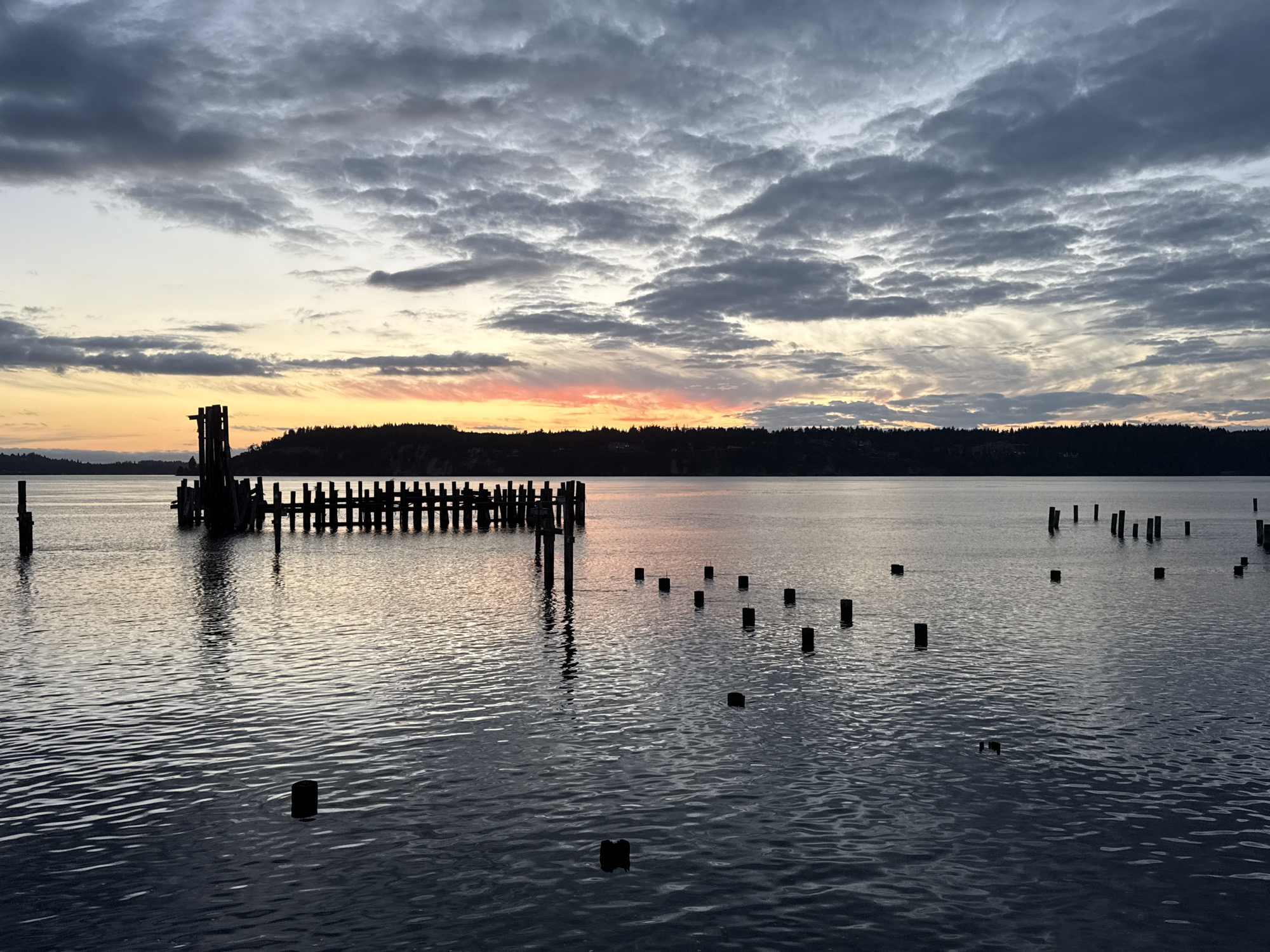 The image depicts a tranquil body of water, likely Commencement Bay in Tacoma, Washington, at dusk. The sky is filled with a dramatic display of clouds, ranging from deep grays to lighter shades, with a warm glow of orange and pink near the horizon, indicating the setting or rising sun. In the foreground, the water is calm, reflecting the muted colors of the sky. Several dark, vertical pilings emerge from the water at varying distances, suggesting the remnants of a pier or dock. To the left, a more substantial, dilapidated wooden structure, also appearing to be a pier, stands in silhouette against the water and sky. The distant shoreline is lined with a dark, dense forest of trees, also silhouetted against the fading light. The overall atmosphere is serene and somewhat melancholic, capturing the quiet beauty of a coastal landscape at twilight. There are no people or animals visible in the frame.