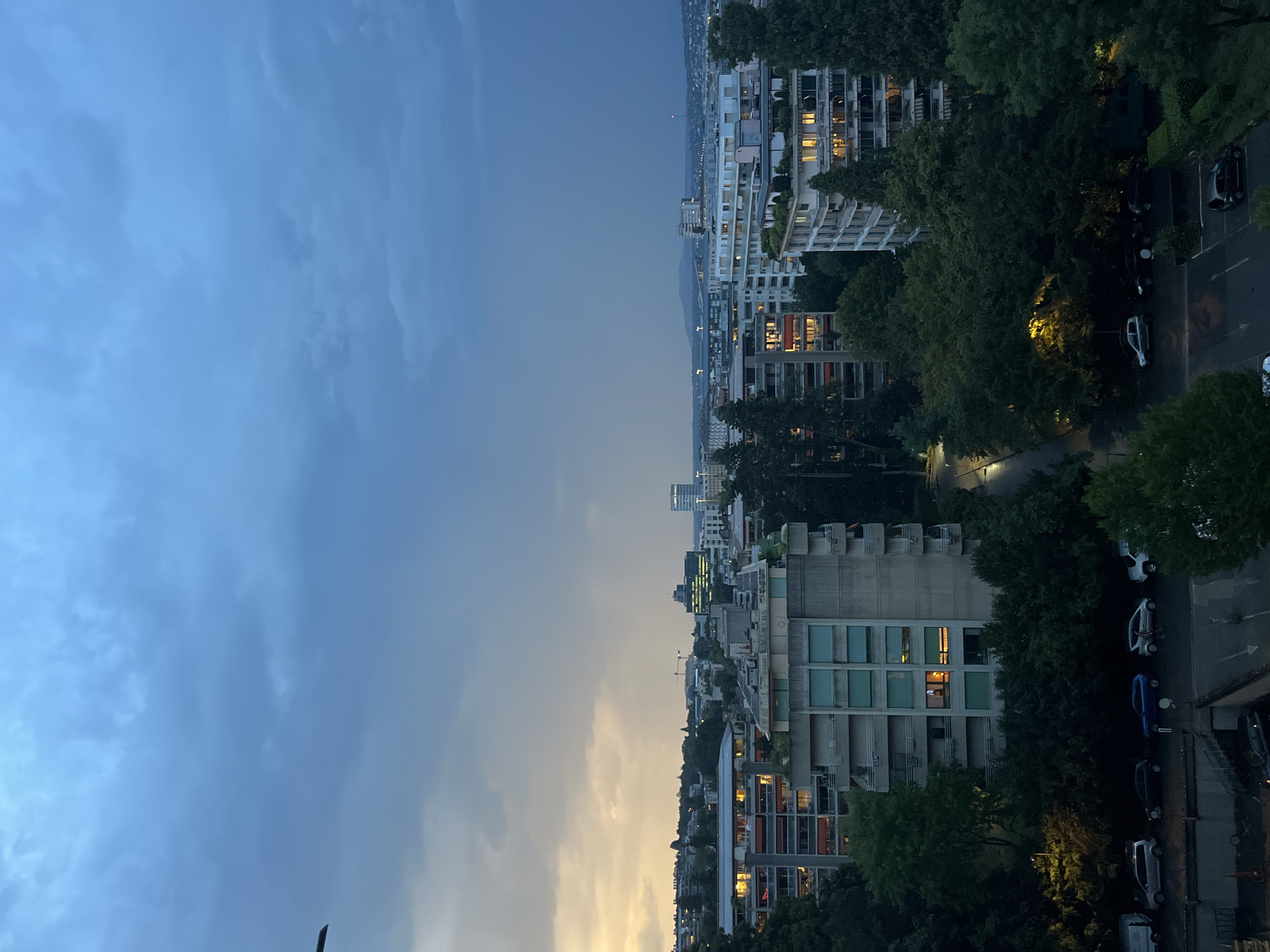 The image shows an aerial view of a city skyline at dusk, likely in Geneva, Switzerland, due to the distinct architectural style and vegetation. The sky is predominantly dark blue with heavy cloud cover, transitioning to a lighter, yellowish hue on the left side, indicating the last rays of sunset. Several multi-story apartment buildings, characterized by their concrete facades and balconies, are visible throughout the scene. Some buildings have illuminated windows, suggesting inhabitants are present. Dense green trees are interspersed among the buildings, creating a contrast between urban development and natural elements. In the foreground, a street is visible with parked cars of various colors and models. A lamppost illuminates a section of the trees. The overall atmosphere is calm and residential, captured during the transition from day to night.