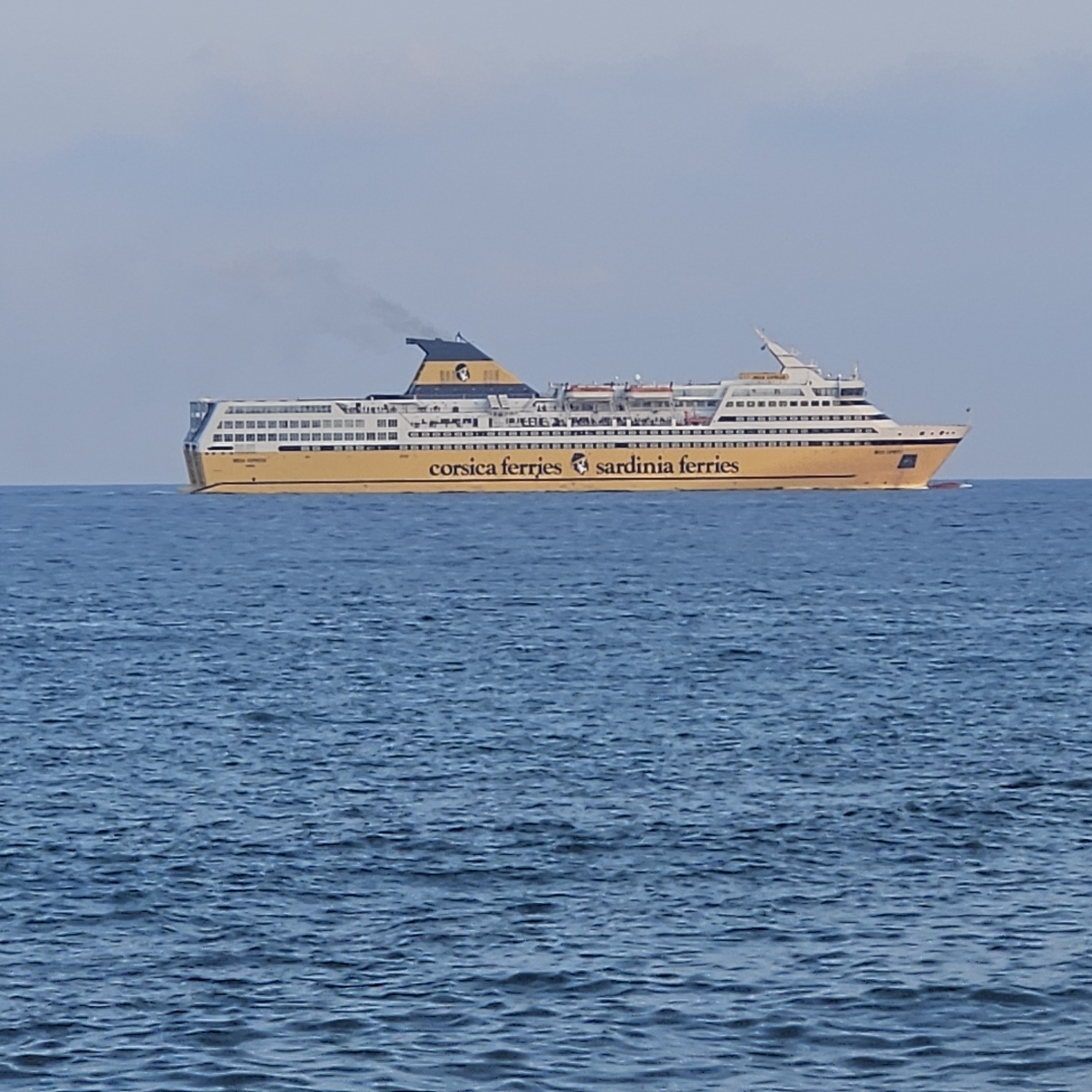 A large yellow ferry boat named "Mega Express" is sailing on a calm blue sea under a hazy sky. The ferry has "Corsica Ferries Sardinia Ferries" written on its side. The ship is mostly white with yellow accents and a distinctive blue and yellow funnel emitting a plume of smoke. Several decks with rows of windows are visible. The water has a slight ripple. The scene is likely a ferry port or along a shipping route near Savona, Italy, given the presence of a large passenger ferry.