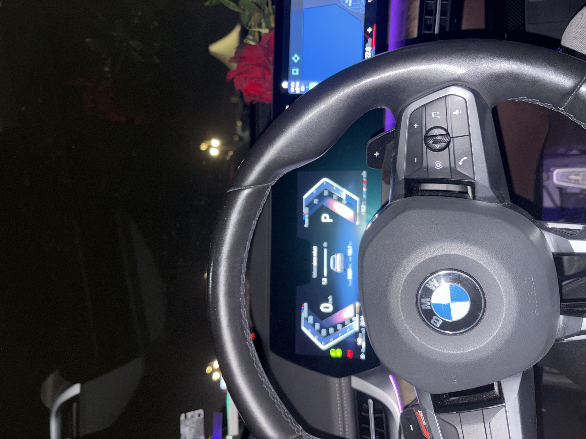 The image shows the interior of a BMW car, specifically the steering wheel and dashboard. The BMW logo is prominently displayed on the steering wheel. The digital dashboard screen is active, showing vehicle information. Outside the car window, a dark, reflective surface is visible, with faint lights suggesting it's nighttime. There is a red rose. There are no people visible in the image. The location in Varna, Bulgaria is not discernible from the visual data provided.