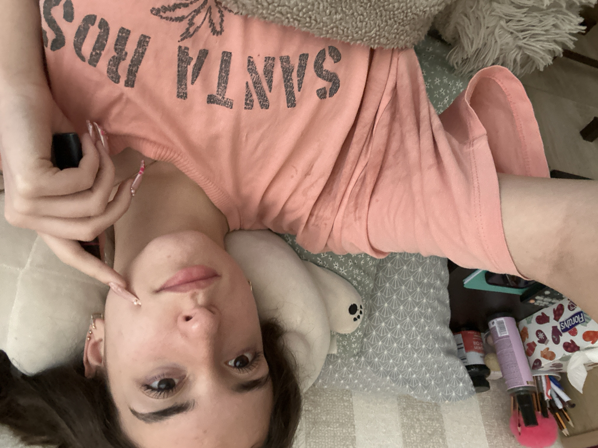The image shows a young woman lying on a bed, taking a selfie. She is wearing a pink t-shirt with the text "SANTA ROSA" printed on it. She has long, dark hair and is wearing earrings. Her right hand is raised to her face, and she is wearing long, decorated fingernails. Behind her, on the bed, is a grey pillow with a white pattern and a white stuffed animal. To the left of the bed, on a nightstand, are several items including a box of tissues, a bottle of spray, and two pill bottles. The background appears to be a wall with a striped pattern. The scene suggests a casual, indoor setting, likely a bedroom. There is no information about the location in the city of Shumen, Bulgaria.