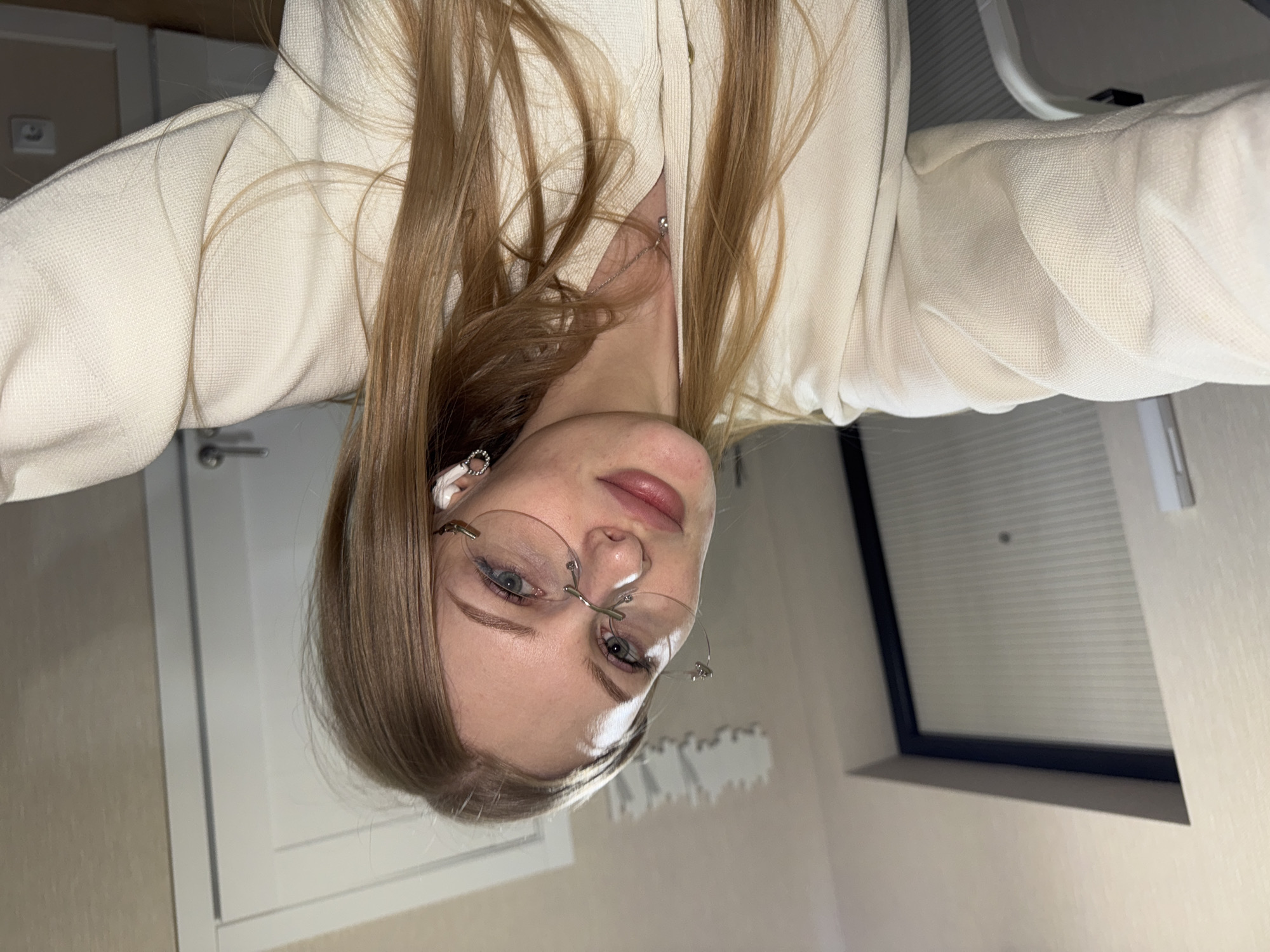 A close-up portrait features a young woman with long, straight, light brown hair flowing down her shoulders. She wears thin, rimless eyeglasses over her light blue or grey eyes, which gaze directly at the viewer. Her lips are a soft pink, and she has subtle eye makeup.
She is dressed in a cream or off-white top, possibly a jacket, made from a textured, woven fabric. A delicate silver chain necklace with a small pendant rests at her collarbone. In her left ear, she wears a white AirPod earbud and a small silver hoop earring.
The setting is indoors, with light-colored walls. A white door with a silver handle is visible to her right. To her left, a window with a black frame and horizontal blinds can be seen. A white light switch or power outlet is on the wall behind her right shoulder. The lighting is soft, suggesting an indoor light source or an overcast day. The woman is posing for the photograph. No specific activity or event is taking place beyond this. There is no visible text in the image. The context for the location is Perm, Russia.