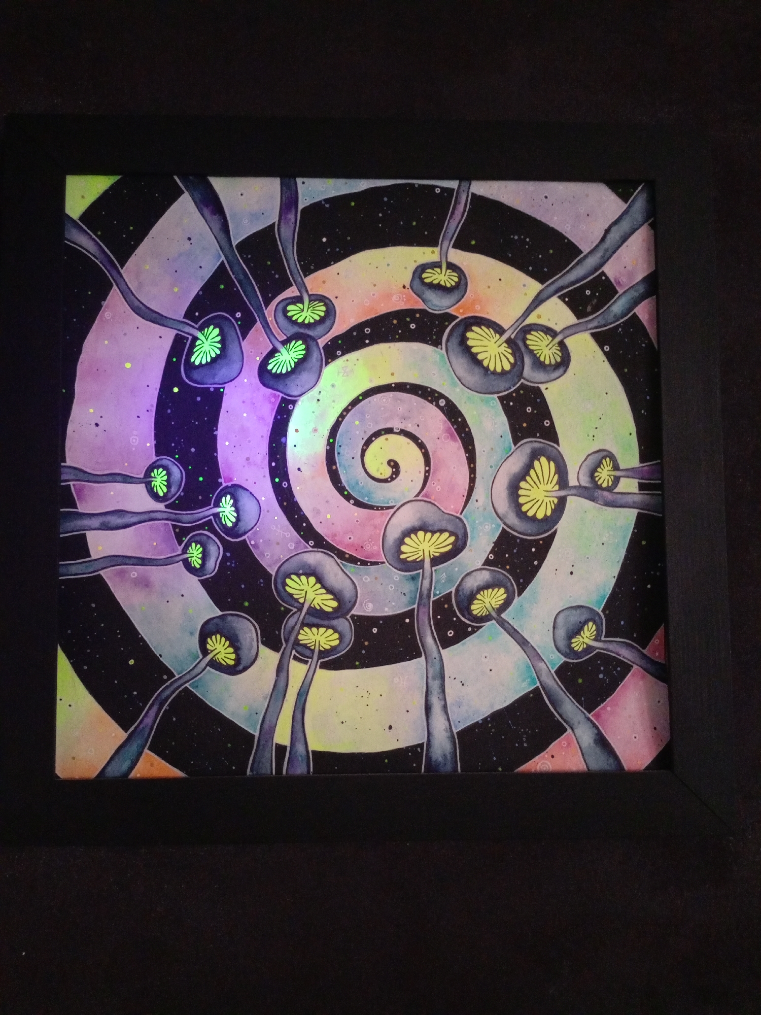 The image shows a framed painting with a psychedelic theme. The painting depicts multiple mushroom-like figures with glowing yellow gills against a swirling, rainbow-colored background with black and white stripes. The mushrooms have dark grey caps and stems. The overall impression is that of a surreal and abstract artwork. The painting is set against a dark background, and appears to be illuminated by a light source that is causing some parts of the painting to glow. The location of this painting within the city of Saratov, Russia is not discernible from the image.