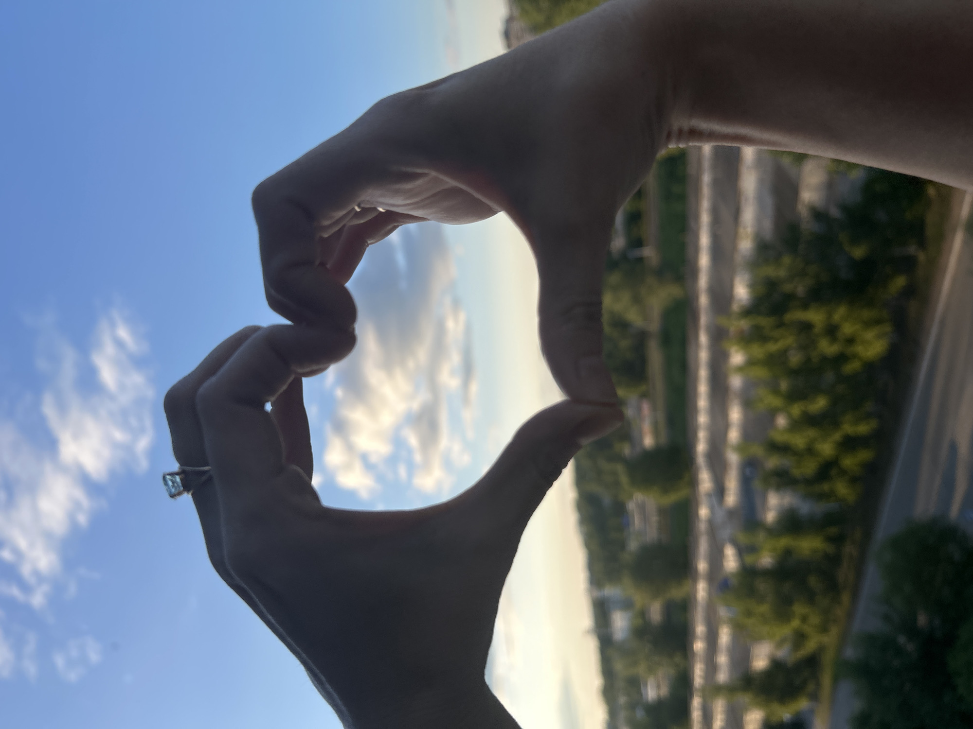 The image shows two hands forming a heart shape against a backdrop of a cloudy sky and a blurred cityscape. On the left hand, a ring with a square-shaped stone is visible. The background includes trees and buildings, suggesting an urban or suburban environment. The lighting indicates it is either sunrise or sunset. No specific location in Chaikovsky, Russia can be identified.