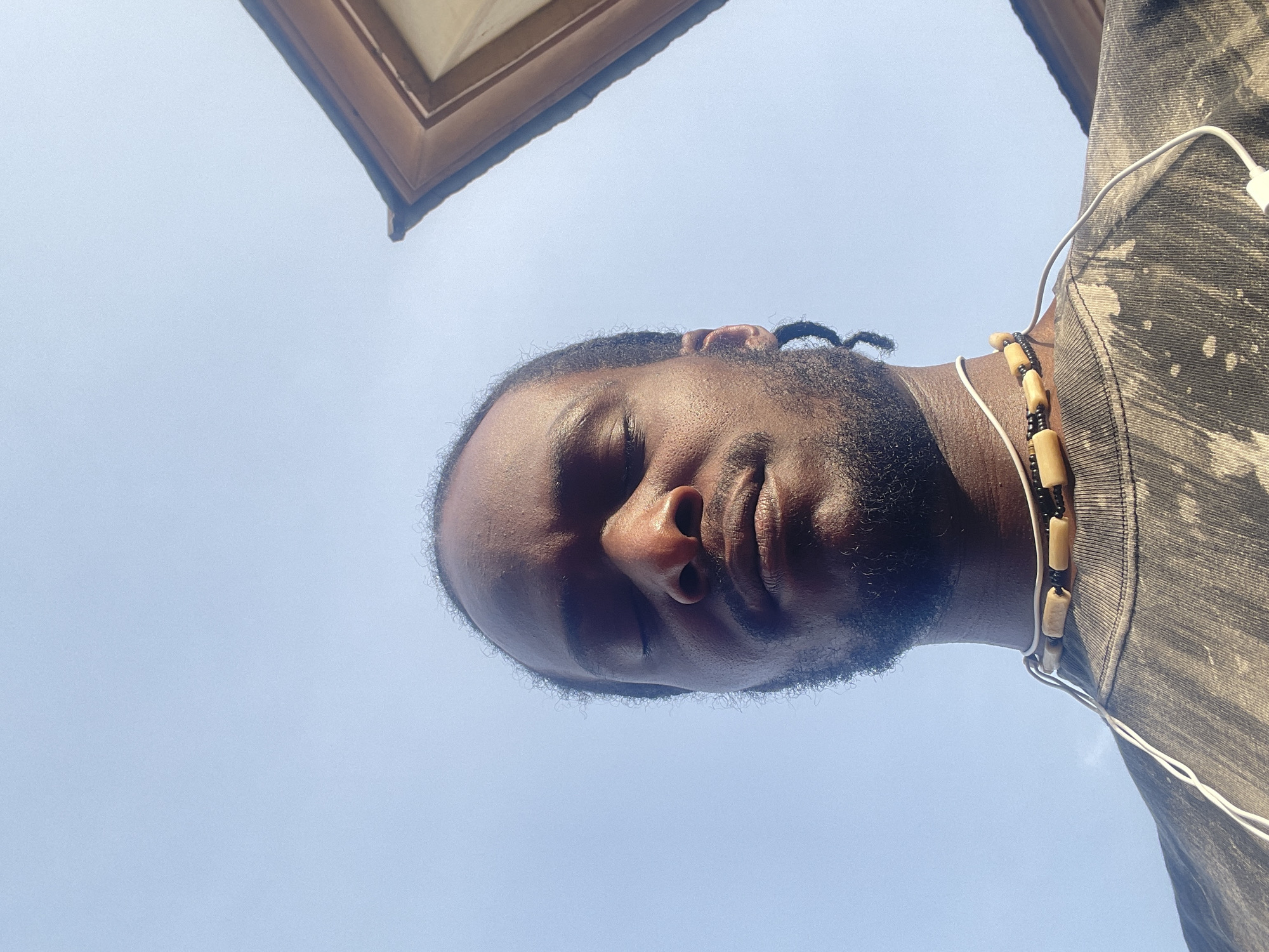 A low-angle shot of a Black male with short hair and a beard, facing downward, presumably taking a selfie. He is wearing a gray shirt and a beaded necklace. An earphone wire is visible near his neck. Above him, a section of a building's structure, possibly a window frame, is seen against a clear, light blue sky. The location is unspecified, and there is no indication of it being Jos, Nigeria.