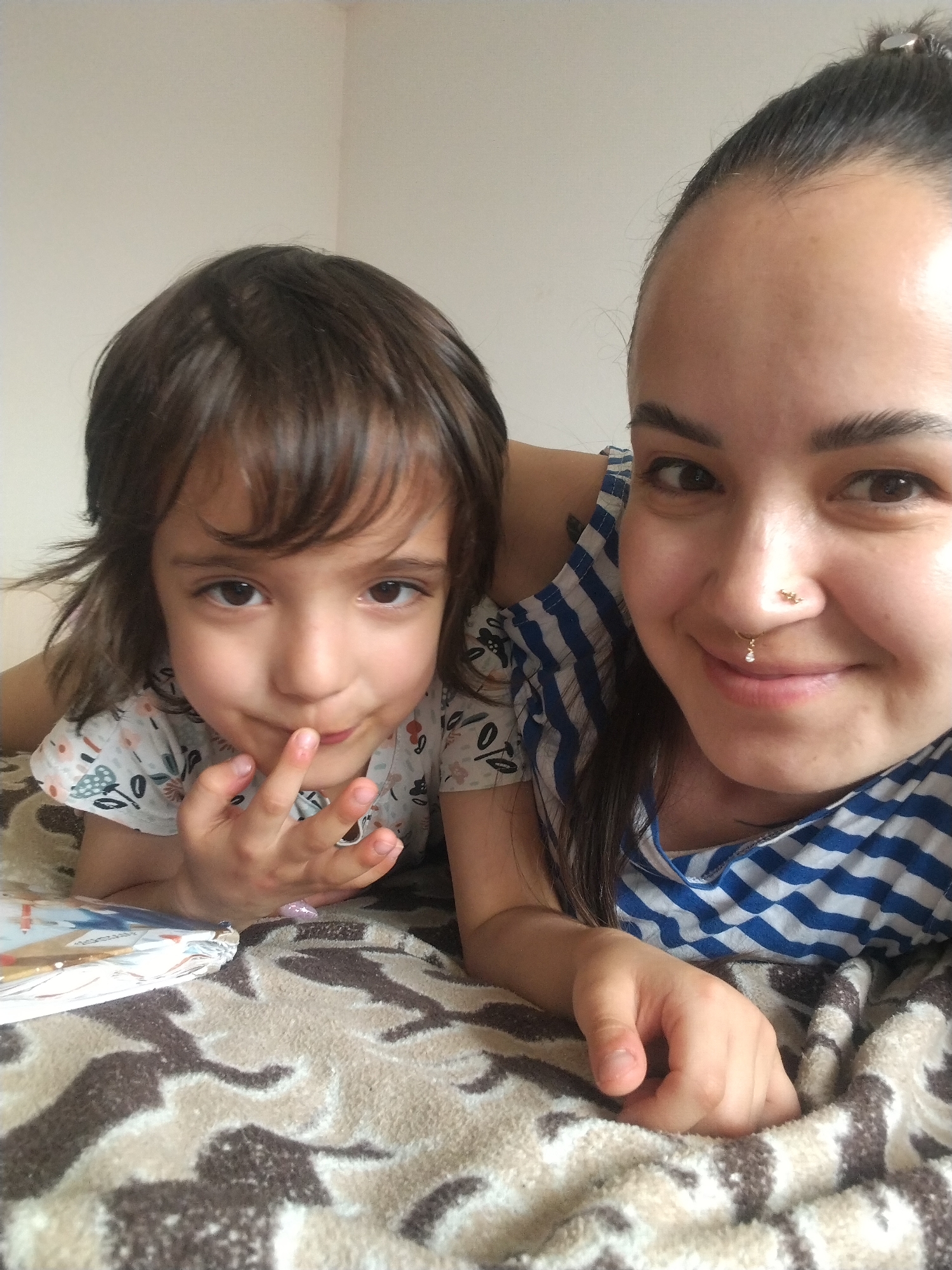 A close-up shot depicts a woman and a child, likely a mother and daughter, lying on a bed covered with a beige and brown patterned blanket. The child, with dark hair and eyes, is positioned to the left, looking directly at the camera with her fingers near her lips. To the right, the woman with her hair pulled back smiles into the camera, wearing a blue and white striped top. She has multiple piercings on her nose. A book with visible pages can be seen slightly obscured to the left of the child. The background is a plain, light-colored wall. The image was probably taken indoors. There is no information in this image about Noviy, Russia.