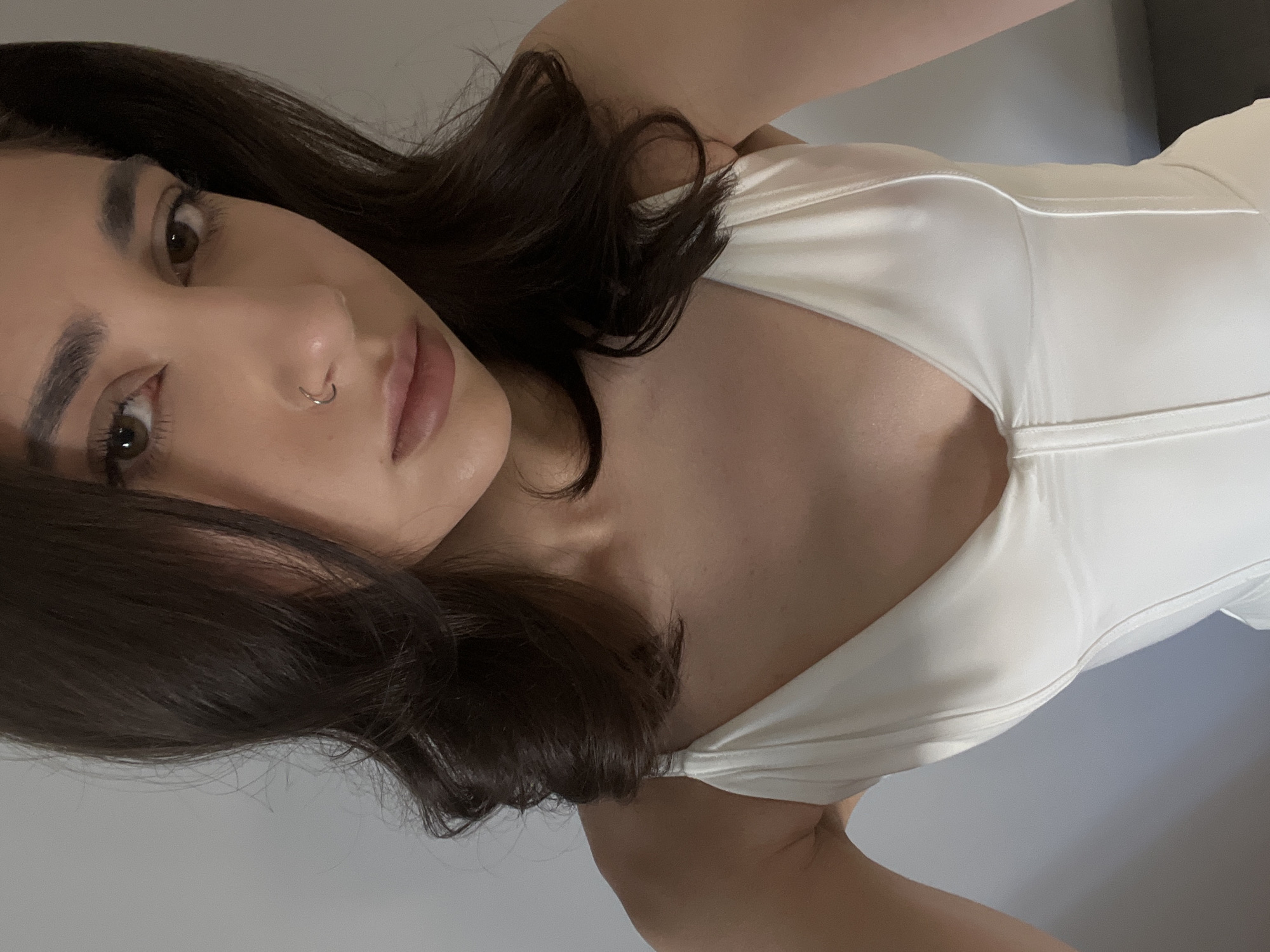 The image is a close-up, head and shoulders selfie of a young woman with dark hair and brown eyes, wearing a white top. She has a nose ring. The background is a plain, light-colored wall. The image does not provide any information about the location being Ternopil, Ukraine.