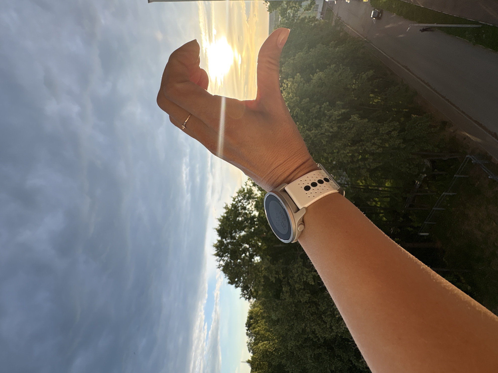 The media file is a vertical, eye-level shot of a person's forearm and hand against a backdrop of a cloudy sky and distant trees, with a road visible in the background. The sun is setting or rising, casting a warm glow. The person is wearing a white smartwatch with a black patterned strap on their wrist. Their thumb is extended upwards, forming a "thumbs up" gesture. A delicate ring is visible on their ring finger.
The scene appears to be outdoors, possibly from a high vantage point like a balcony or window, given the view of the road and trees below. The sky is filled with dramatic clouds, suggesting a dynamic weather system or the transition of day to night. The light indicates it is either early morning or late evening. The overall mood is positive, conveyed by the "thumbs up" gesture. The visible text on the smartwatch face is "20:55" and "5924". The location context provided is Chaikovsky, Russia.