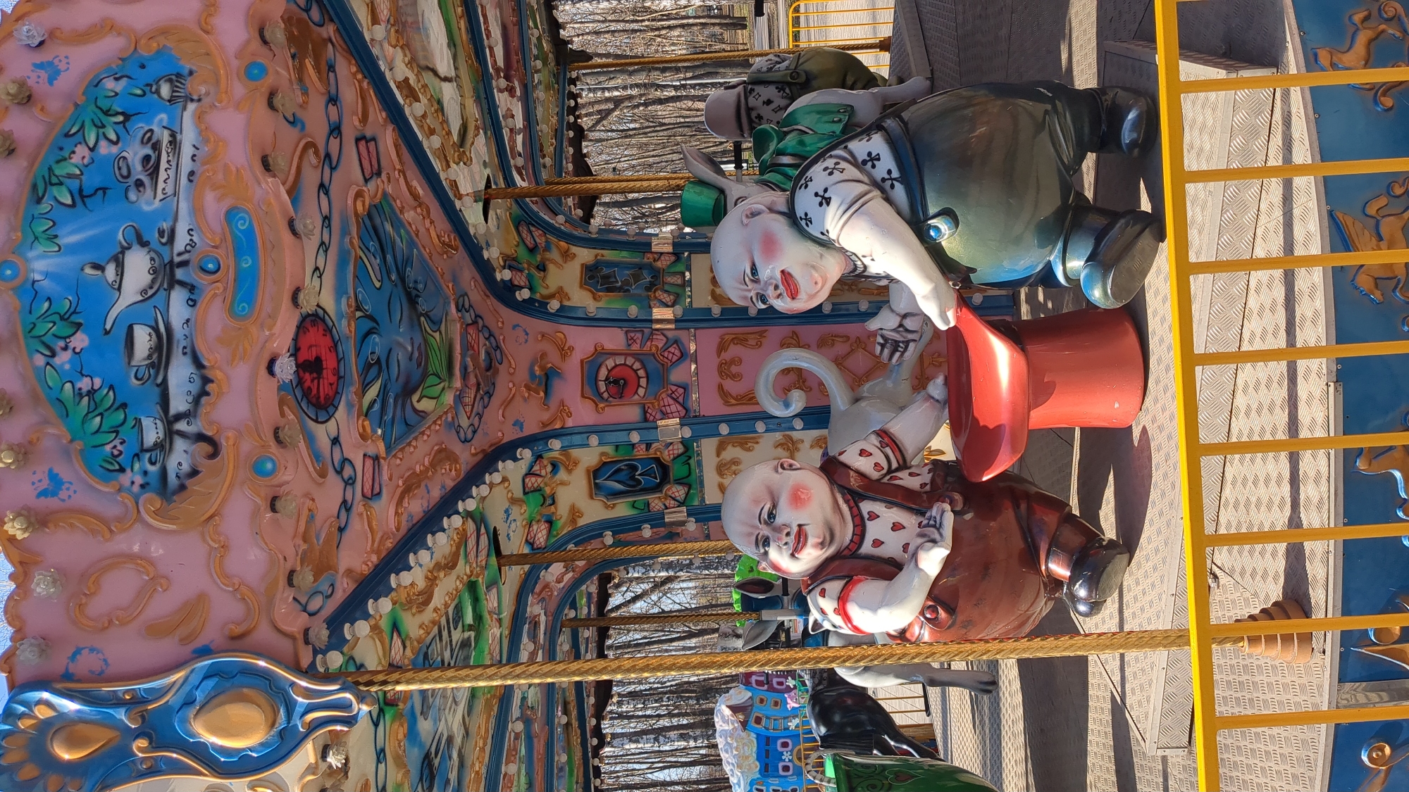 The image displays a brightly lit carousel ride, likely situated in an amusement park or fairground in Ulyanovsk, Russia. The scene is illuminated by strong daylight, casting clear shadows on the textured metal platform floor.
The main subjects are several large, stylized figures on the carousel, reminiscent of characters from "Alice in Wonderland." Two prominent figures have pale, exaggerated faces. One wears a green hat and a green and brown outfit, appearing to hold a red top hat. The other, positioned closer to the viewer, is clad in a white shirt adorned with red heart patterns, sporting a slight smile. A third figure with rabbit-like ears is partially visible further back.
The carousel's panels are elaborately decorated with vibrant pink and blue sections outlined in gold. These panels feature intricate artwork including painted scenes of teacups and teapots, stylized chains, and playing card motifs like spades and hearts. A notable detail is a red-faced clock embedded within one of the blue sections. Small ornamental elements such as butterflies and flowers also adorn the carousel. On the right side of the frame, a yellow metal railing or ladder is visible. In the background, beyond the carousel structure, the trunks of bare trees can be seen, indicating an outdoor setting. There is no discernible text visible on the ride or the figures.