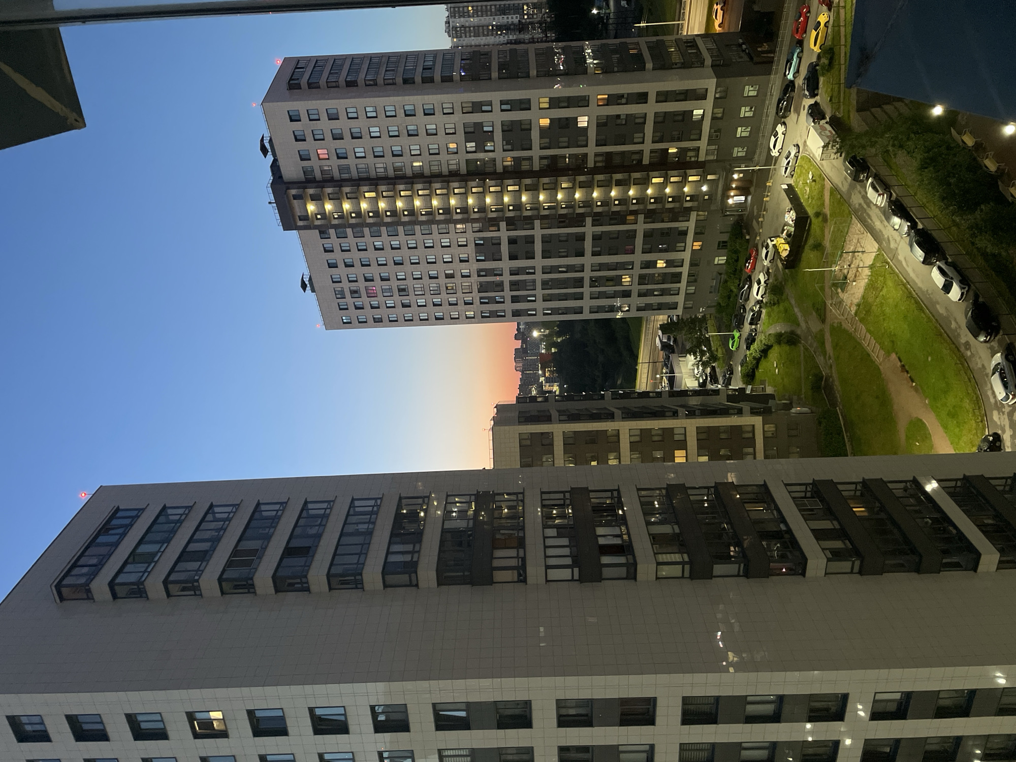The image depicts an aerial view of a residential area in Sinyavino, Russia, at dusk. Two tall apartment buildings dominate the foreground. The building on the left is lighter in color, with a facade of pale tiles and dark-framed windows. The building on the right has a more complex facade, featuring both light-colored panels and darker sections, with numerous windows illuminated from within, indicating occupancy. Several smaller apartment buildings and structures are visible in the background, receding into the distance.
Below the buildings, a street lined with parked cars can be seen, with vehicles of various colors and models, including sedans and SUVs. Some cars are parked on the side of the road, while others are in designated parking areas. A small patch of green space, possibly a park or courtyard, is situated between the buildings and the street, with a paved path winding through it. The sky is a gradient of deep blue at the top, transitioning to warmer hues of orange and pink near the horizon, characteristic of twilight. The overall scene conveys a sense of urban living and the transition from day to night.