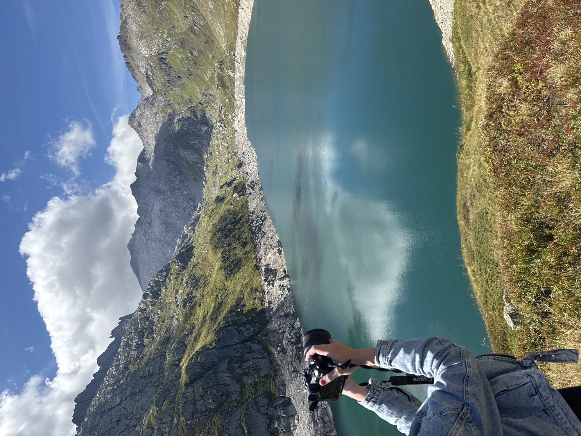 In Airolo, Switzerland, a person in a denim jacket holds a camera, capturing a vast, turquoise lake surrounded by rugged, rocky mountains. The sky is a clear blue with scattered white clouds. The terrain leading down to the lake is covered in dry grass and sparse vegetation. The person is positioned on a grassy slope, looking towards the expansive mountainous landscape and the water.