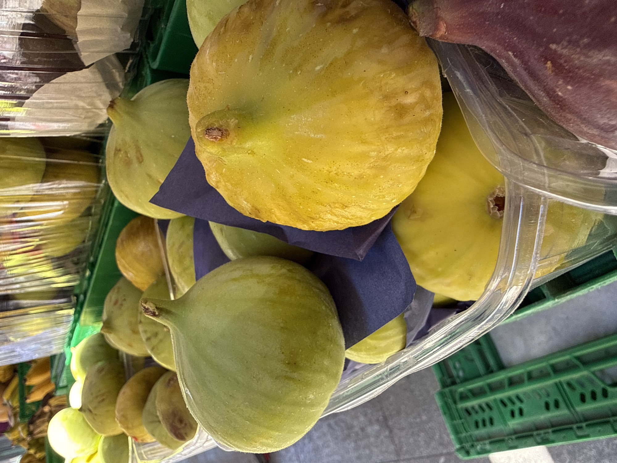 A close-up, eye-level, outdoor shot shows a plastic container filled with fresh figs at a market stall. The figs are of various colors, including green and yellow, and appear plump and ripe. The container is transparent, allowing a clear view of the fruit. In the background, more figs are visible in similar containers, as well as green plastic crates. The scene is illuminated by natural sunlight, casting subtle shadows and highlighting the texture of the figs. The overall impression is one of a bountiful harvest at a local market. This image was captured at a fruit stand in Kadıköy, Istanbul, Turkey.