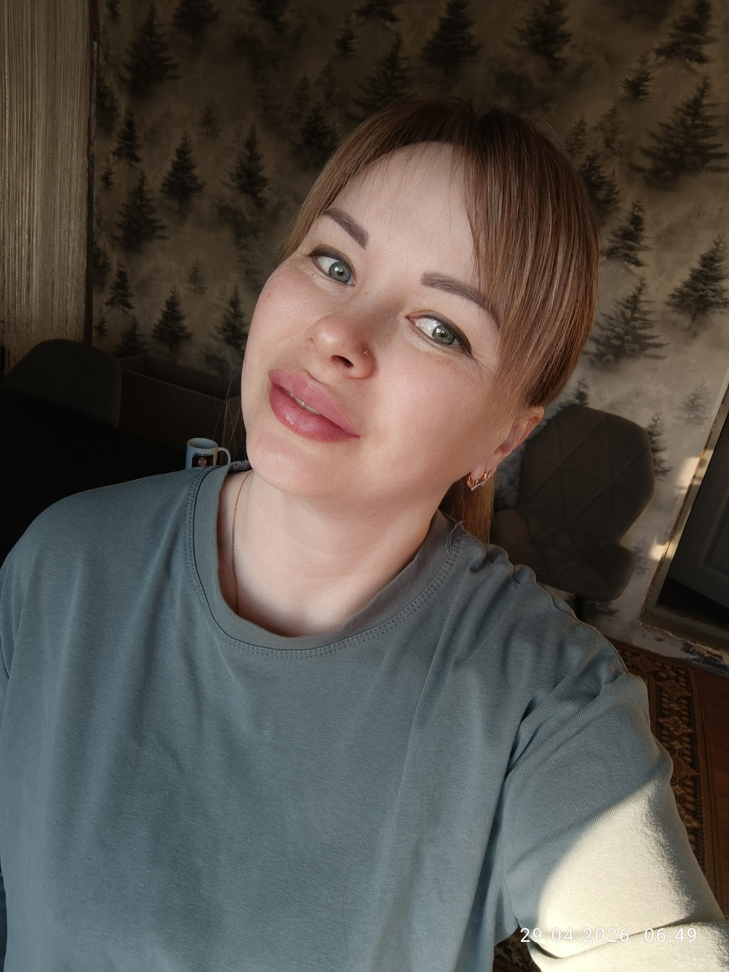 A woman takes a selfie indoors in Chaikovsky, Russia, during the early morning. She has light brown hair with bangs, green eyes, light makeup, a small nose piercing on her right nostril, and delicate gold hoop earrings. She wears a relaxed-fit, long-sleeved teal-grey t-shirt and a thin gold chain necklace. She is looking directly at the camera with a subtle smile.
The background features wallpaper with a repeating pattern of dark, silhouetted pine trees against a lighter, textured brown/grey background. To her left, part of a vertical textured surface, possibly a curtain or wall hanging, is visible. Behind her, a dark chair with a geometric pattern on its back is partially obscured. A white mug with a blue rim and an image (possibly of a person) on it is visible behind her left shoulder. A patterned rug is visible in the bottom right corner.
Strong natural light streams in from the left, creating bright highlights on her face, hair, and the left side of her body, and casting distinct shadows. The time and date "29.04.2026 06:49" are stamped in the bottom right corner of the image, confirming the early morning setting and sunny conditions.