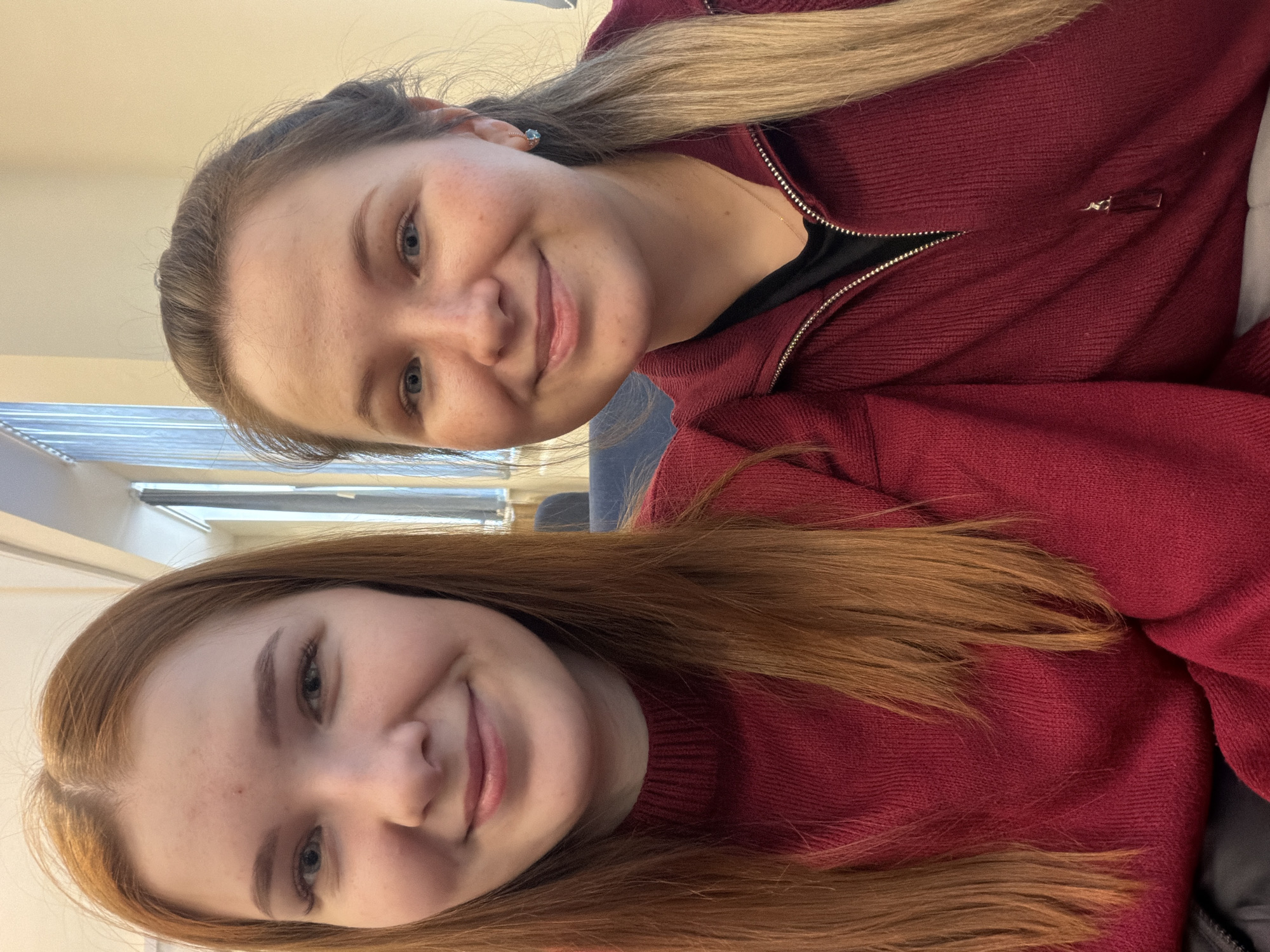 Two young women are depicted in an indoor portrait, smiling warmly towards the camera. The image is oriented sideways, with their heads tilted towards the top.
The woman in the foreground has long, reddish-brown hair flowing over her shoulders and is wearing a deep red, possibly knitted, sweater. She has light-colored eyes and a gentle smile.
Behind her, positioned slightly to her right, is another woman with long blonde hair. She is wearing a dark red or maroon zippered jacket over a black top, and a small, light blue stud earring is visible in her left ear. She also has light-colored eyes and a broad, friendly smile.
The background suggests an indoor setting, with a light-colored ceiling or wall and what appears to be a window with blinds or louvers visible. The lighting is soft and even, typical of an interior space. The subjects' expressions convey happiness and friendliness. There is no discernible text within the image. The scene is informal, possibly a casual gathering or a shared moment. No specific visual cues confirm the location as Chaikovsky, Russia, other than the provided context.