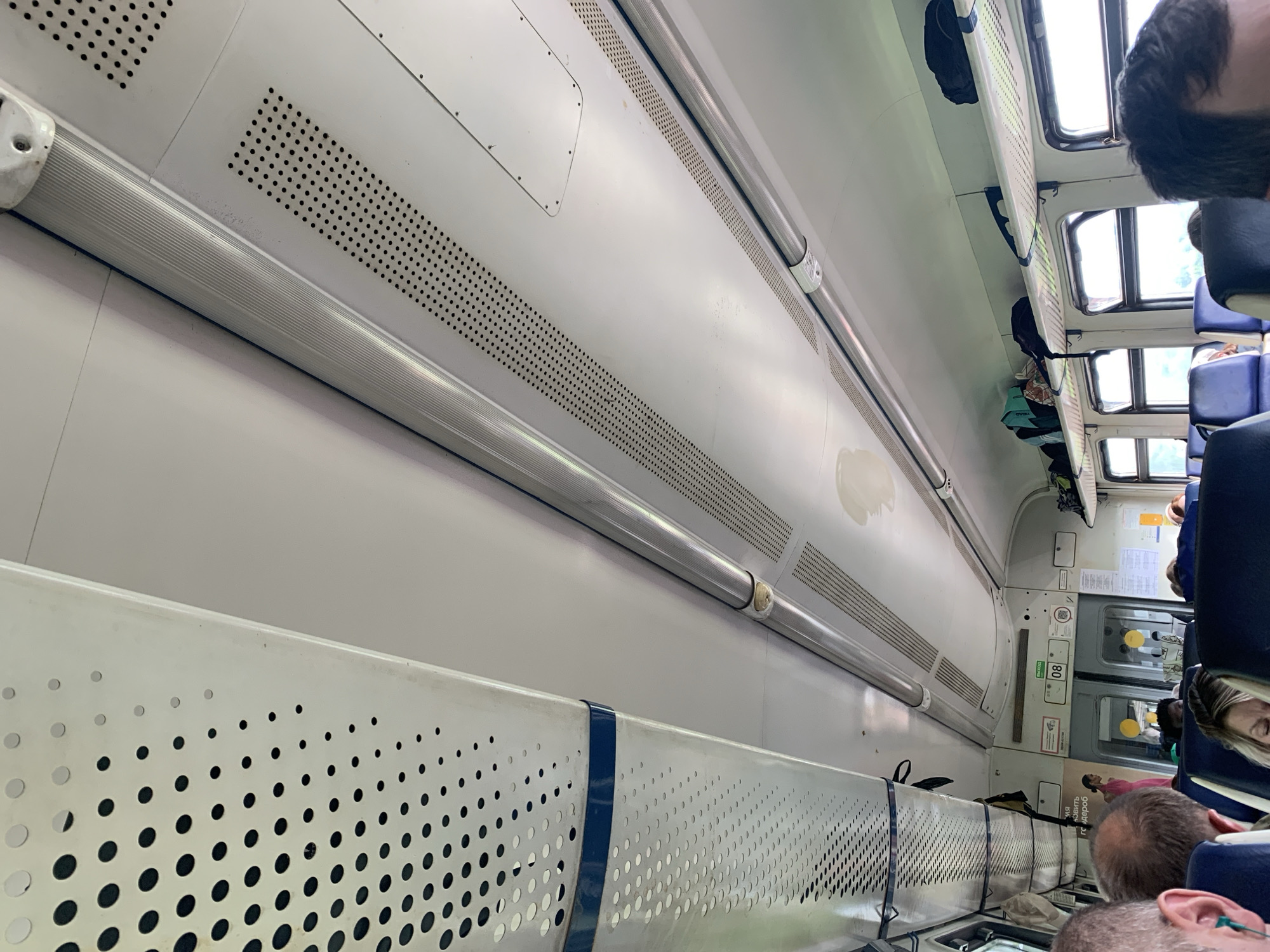 The image shows the interior of a train car, looking upwards towards the ceiling. The ceiling is white and features several light fixtures and ventilation grilles. In the foreground, the heads and shoulders of several passengers are visible, indicating they are seated. On the right side of the image, luggage is stored on an overhead shelf. The windows of the train are visible, revealing a blurry outdoor scene with greenery, suggesting the train is in motion and possibly passing through a rural or suburban area. The overall impression is that of a typical train interior. The location is identified as Kungur, Russia.