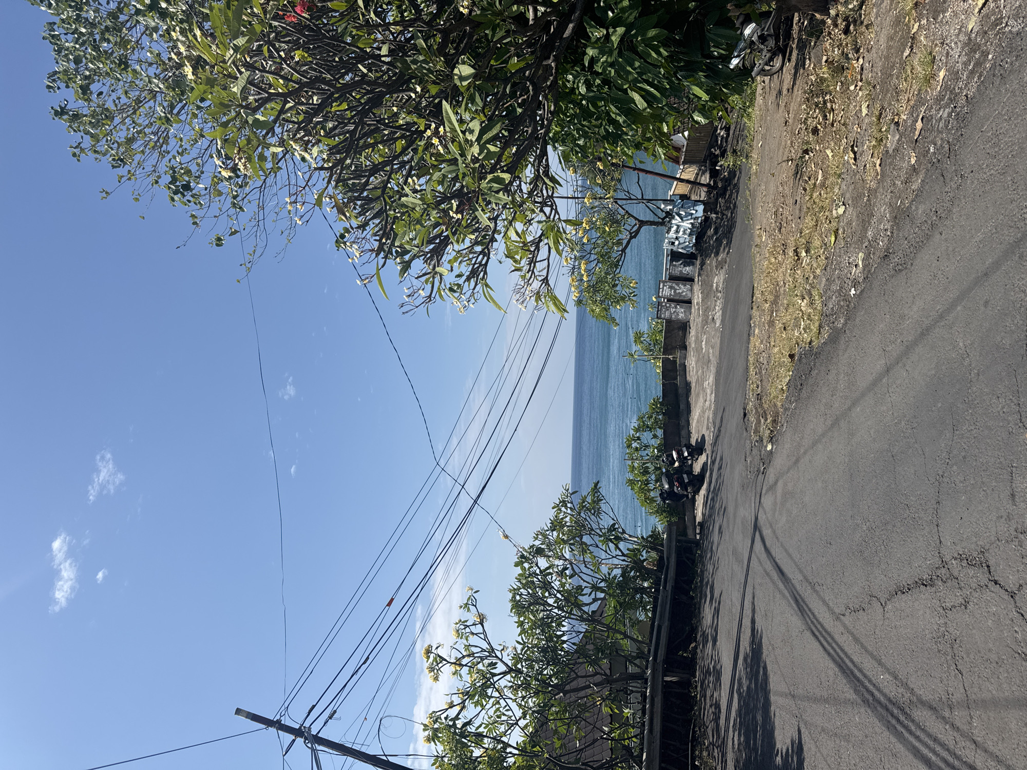 A paved road descends towards a body of water under a clear blue sky in Abang, Indonesia. Trees with green leaves and some flowering branches flank the road. Electrical wires crisscross the sky, supported by a utility pole on the left. Two motorcycles are parked near the edge of the road, and further down, several chalkboards are visible against a wall. The ocean stretches to the horizon, with a slight chop on its surface. The ground beside the road is a mix of dry grass, dirt, and fallen leaves.