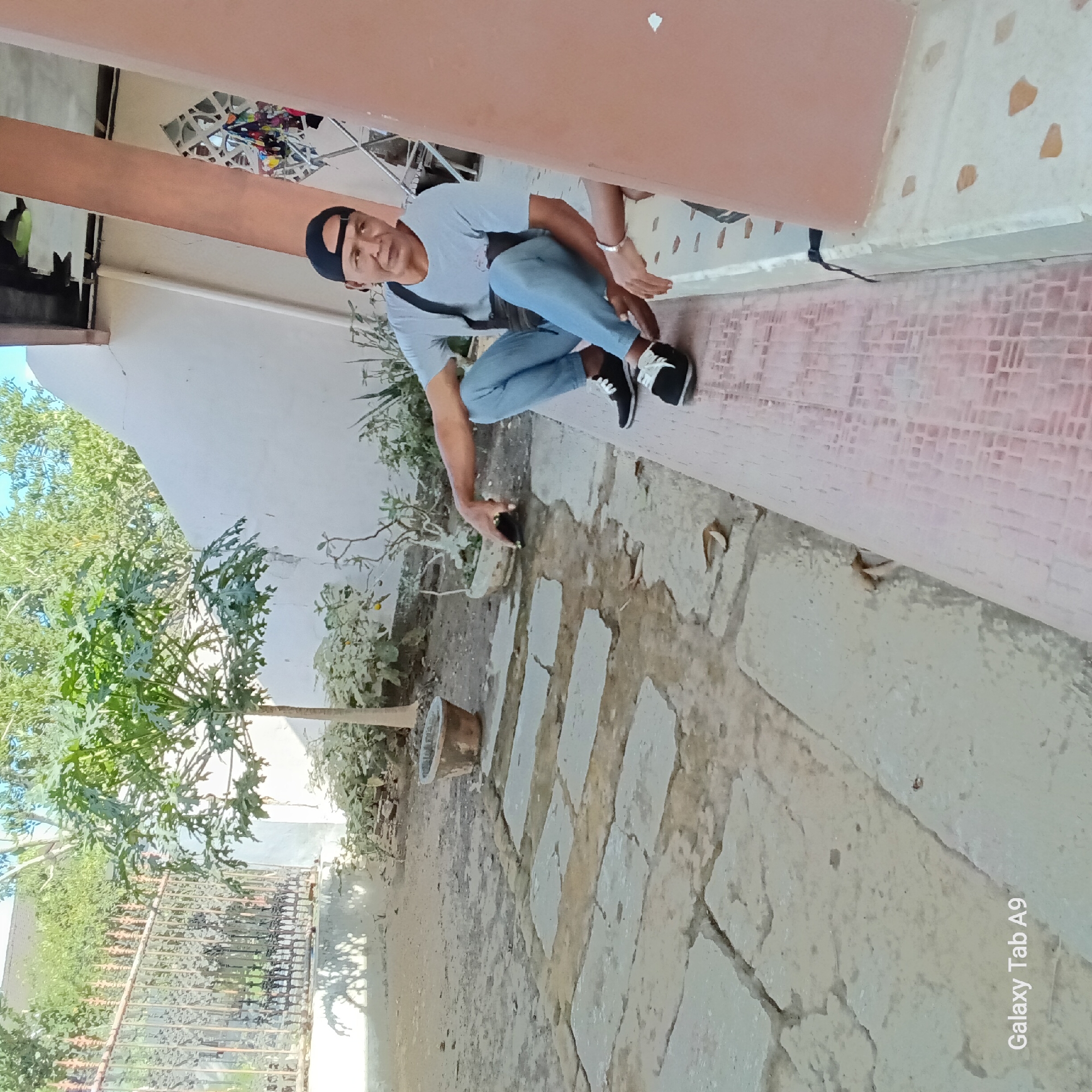 In Sambirejo, Indonesia, a man is sitting on a raised tiled platform next to a pink column. He is wearing a light blue t-shirt, blue jeans, a black baseball cap, and black and white sneakers. He has a black bag slung across his chest. His left arm is resting on his leg and his right hand is reaching towards a black object on the ground. Another person's arm and hand are visible in the lower right corner of the image. The background shows a gravel courtyard with a stone pathway, a tree, potted plants, and a metal fence. Sunlight casts shadows on the ground. The wall behind the man appears to be painted white.