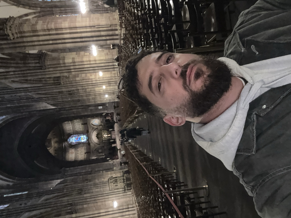 A male individual with a beard and mustache, wearing a light grey hooded sweatshirt under a dark jacket, occupies the foreground, looking directly at the camera in a selfie perspective. He is positioned inside the Cathédrale Notre-Dame de Strasbourg, France. The background reveals the expansive Gothic interior of the cathedral, characterized by towering stone columns and ribbed vaulted ceilings extending into the distance. Rows of dark wooden chairs line both sides of a central aisle, which is delineated by low barriers. Several indistinct figures are visible walking down the aisle towards the far end, and others are seated among the chairs. A large stained-glass window, predominantly blue, is visible at the far end of the nave, illuminating that section. The cathedral interior is moderately lit, showcasing the intricate stone architecture.