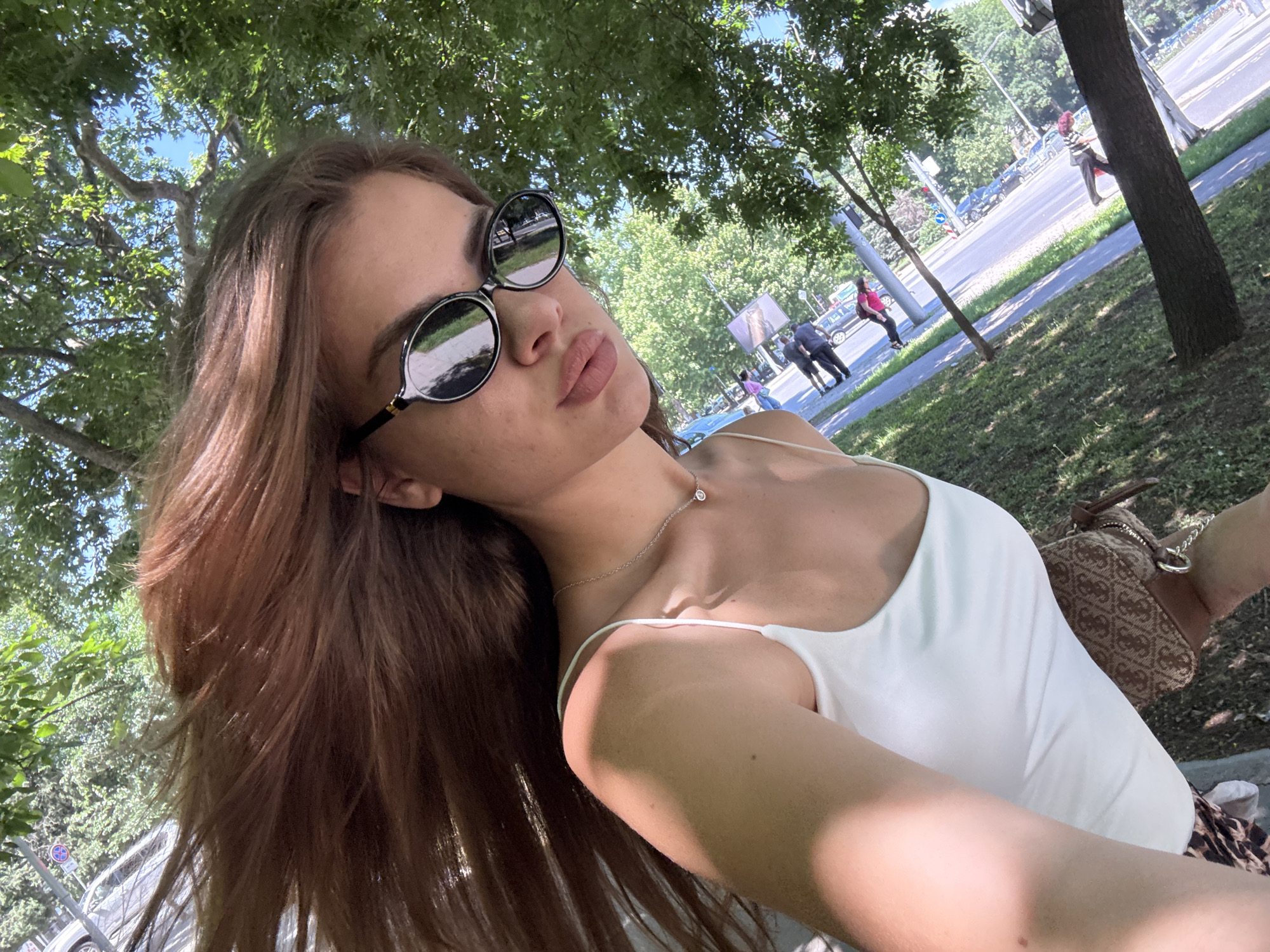 A young woman with long, brown hair and sunglasses is taking a selfie outdoors. She is wearing a white tank top and a necklace. Her hair is blowing in the wind. In the background, there is a street with cars and people walking. The scene appears to be in a park or along a tree-lined street. Based on the architecture visible and the context of the image, this location is likely near the Plovdiv Roman Stadium, a historic landmark in Plovdiv, Bulgaria, characterized by its ancient ruins adjacent to modern city infrastructure.