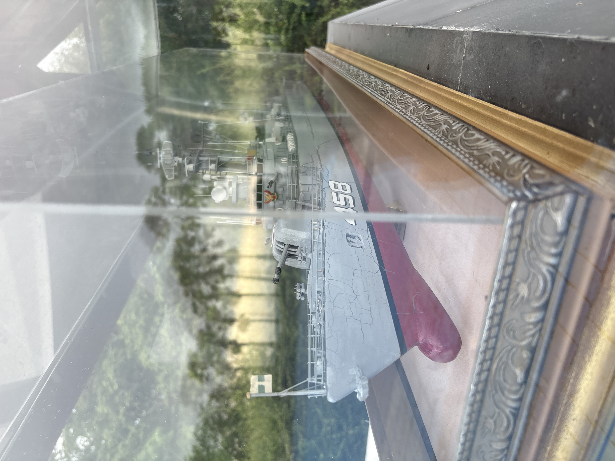 This is a medium-shot, eye-level, outdoor photograph of a detailed model warship displayed inside a clear glass case. The warship is predominantly grey with a red hull. Visible details include cannons, a radar tower, a flag on the bow, and the number "158" painted on its side. The model is placed on a beige surface within the case, which itself is framed by an ornate gold and silver border. The background consists of blurry green foliage and trees, suggesting a natural outdoor setting. The image is taken from a slightly elevated angle, looking down and across the display case. The lighting suggests it is daytime, with some sun glare on the glass case, indicating it is located somewhere in Rawai, Thailand.