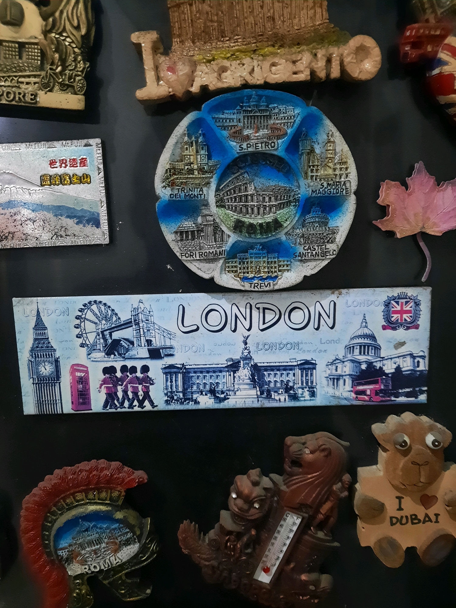 The image is a close-up, overhead shot of a collection of refrigerator magnets displayed on a dark, flat surface. The magnets depict various landmarks and cultural symbols.
Starting from the top left, there is a magnet with a golden structure and the word "PORE" visible. To its right, a larger, beige magnet displays a textured architectural element with the word "CRGENTO" and part of the letter "I" preceding it. To the right of that, a magnet with a red, white, and blue pattern, resembling a Union Jack, is partially visible.
Below the "PORE" magnet is a silver rectangular magnet with Asian characters, including "世界遗产" (World Heritage) and what appears to be a depiction of Mount Fuji.
To the right of the Asian magnet, a circular magnet is divided into sections, each featuring a different Roman landmark. Visible labels include "S. PIETRO," "COLOSSEUM," "FORI ROMANI," "TREVI," "S. MARIA MAGGIORE," and "CASTEL SANT'ANGELO." A light pink, leaf-shaped magnet is situated to the right of this circular magnet.
Below these magnets, a horizontal rectangular magnet is predominantly blue and white, depicting various London landmarks. Prominently featured are Big Ben, the London Eye, Tower Bridge, and several red double-decker buses and guardsmen. The word "LONDON" is written in large, bold letters across the top of this magnet.
In the bottom section of the image, a magnet shaped like a Roman soldier's helmet with a red plume is visible. Inside the helmet, a depiction of Rome, including the Colosseum, is visible with "ROMA" written below it. To the right of the helmet magnet, there are two more magnets. One is a dark brown, decorative object that appears to incorporate a thermometer. The other is a light brown, teddy bear-like figure with the text "I ❤ DUBAI" written on its chest.
The scene appears to be a collection of souvenirs, likely on a refrigerator. There are no people present in the image, and the objects are static. The background is dark and uniform, providing no environmental context for a specific location. The image does not contain any indicators that would suggest a location within East Jakarta, Indonesia.