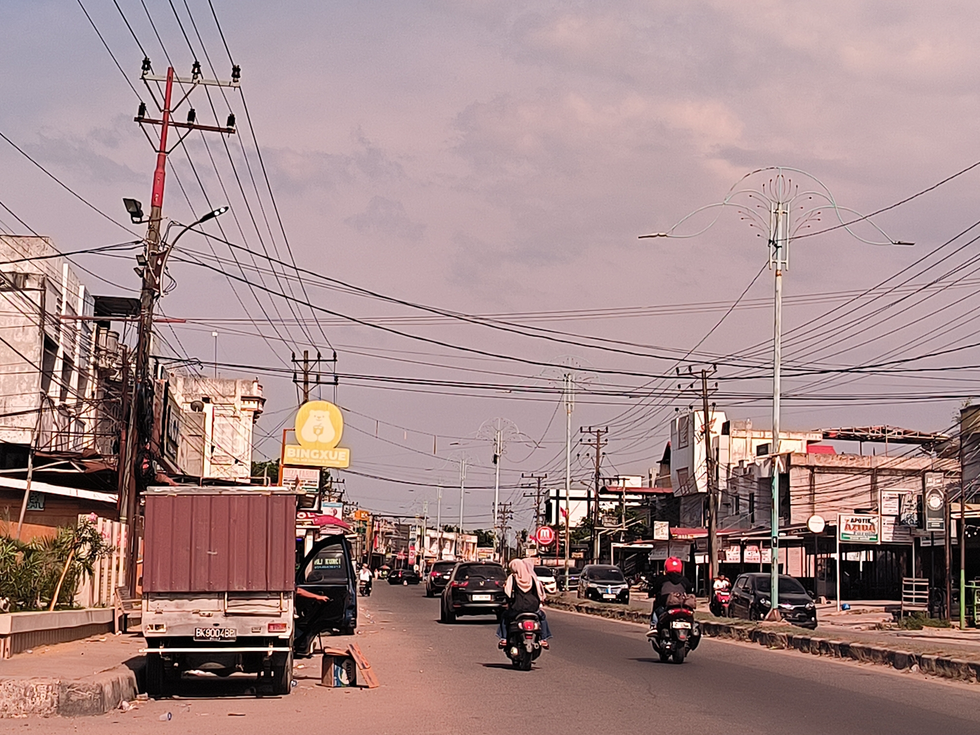 The image captures a street scene in Titibaro, Indonesia. A truck with a maroon container body is parked on the left side of the road, its license plate visible. A yellow "BINGXUE" sign is seen in the background. The road has light vehicular traffic, with cars and motorcycles moving in both directions. Two motorcyclists are in the foreground, one with a pillion rider wearing a hijab. Overhead power lines are strung between utility poles that line the street. Buildings are visible on either side of the road. On the right, a sign for "APOTIK AZIDA" is seen. The sky is overcast.