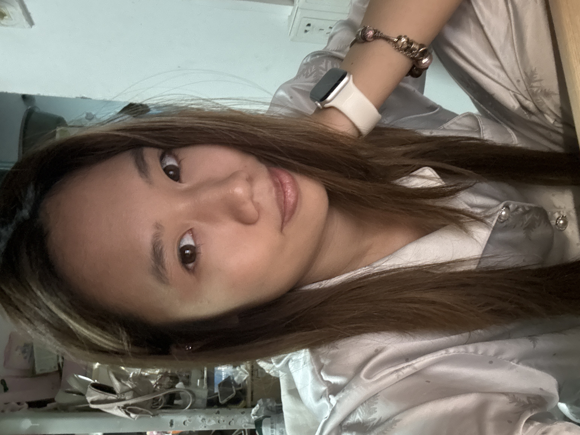 A woman with long, light brown hair and a white smartwatch is posing indoors. She is wearing a light-colored satiny top. The immediate background is cluttered with items on a shelf, including what appears to be packaged goods. Above her, there is a white wall with an electrical outlet. There is no visible evidence to confirm the location is Nan'an District, China.