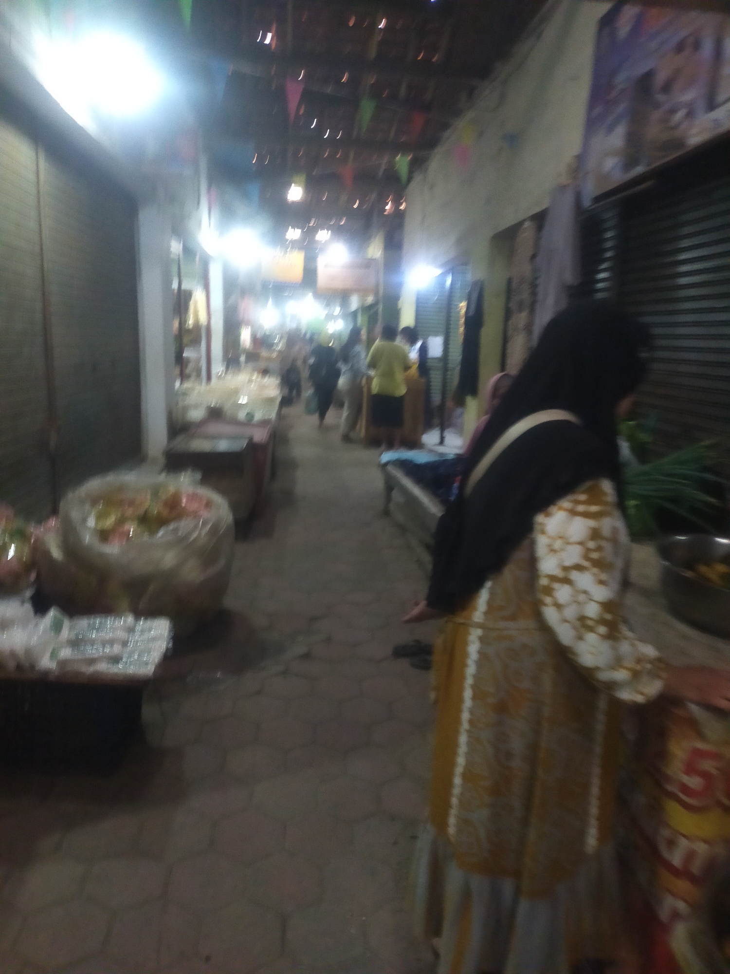 The image depicts a busy outdoor market in Trayeman, Indonesia, characterized by its narrow alleyway and overhead string lights. The market appears to be in operation, with several individuals present and various goods displayed for sale.
In the foreground, a woman wearing a yellow and white patterned dress and a black hijab is visible from the chest down, facing away from the camera. Her right arm is extended slightly. To her left, a large, clear plastic bag filled with colorful snacks or food items is prominent on a table. Adjacent to this, to the left of the bag, are several smaller packages wrapped in clear plastic, possibly more food items or products.
Further down the alleyway, other market stalls and shoppers can be observed. To the left, a closed metal shutter suggests a shop or stall. In the background, more stalls are present, with a variety of items and people milling about. The floor of the alleyway is paved with hexagonal tiles. The overall lighting suggests it might be evening or a dimly lit indoor market. The atmosphere is one of a bustling local marketplace.