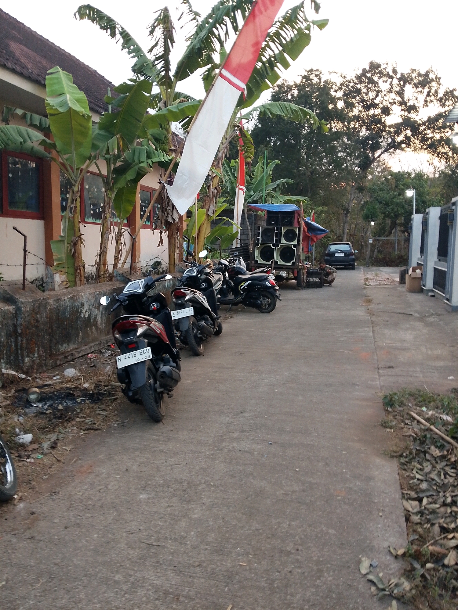 The image shows a street in the city of Karangsari, Indonesia, with several parked motorcycles on the right side of the street. A building with a brown roof and windows is visible on the left side of the image, with banana trees growing in front of it. A large sound system with multiple speakers is set up on a cart in the middle of the street, with a person kneeling next to it. A dark-colored car is parked further down the street. The ground is paved with concrete, and there are fallen leaves and debris on the side of the road. A red and white Indonesian flag is visible on a pole in the background. The scene appears to be outdoors, with natural lighting.