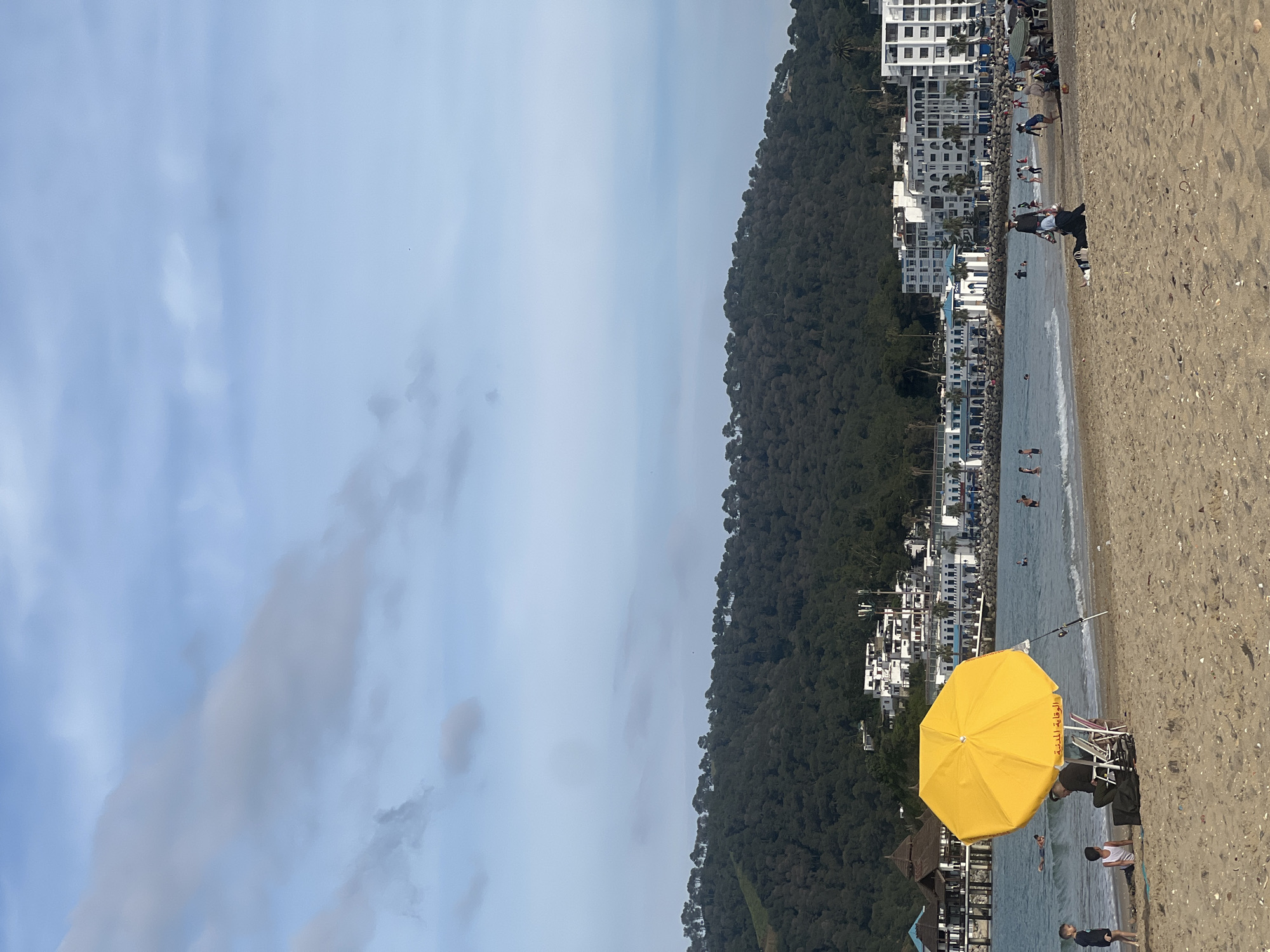 The image depicts a beach scene in M'Diq, Morocco. A yellow beach umbrella is positioned in the foreground on the sandy shore. People are present on the beach, some in the water and others on the sand. Behind the beach is a hillside covered in trees, and white buildings can be seen interspersed with the trees and along the coastline. The sky is overcast with clouds.