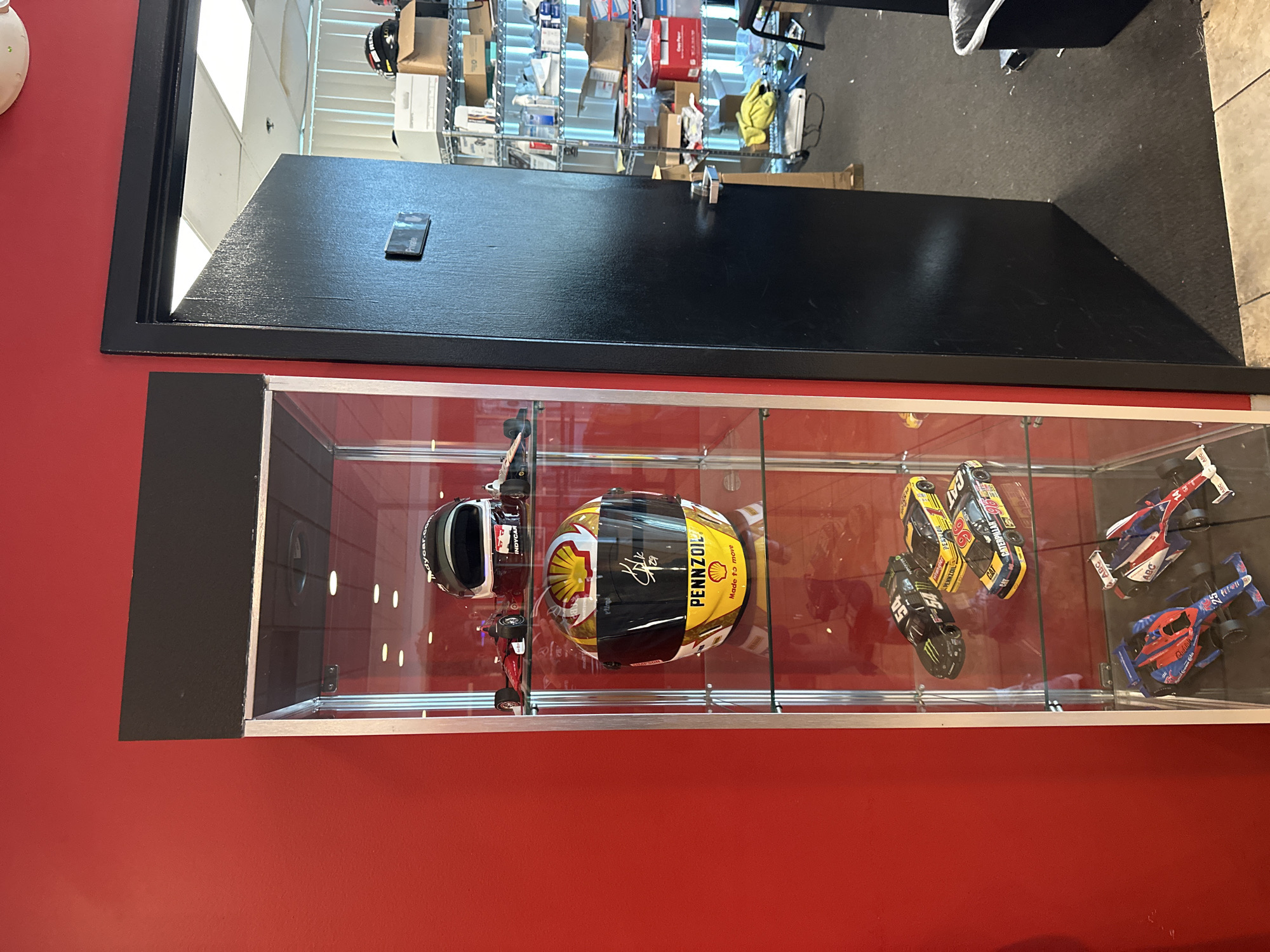 A display case is mounted on a red wall. The display case contains two race car helmets and four model race cars. The helmets are a black and white helmet and a yellow and black helmet with Pennzoil logos. The model race cars are various colors. The display case is in Orient Park, United States of America.