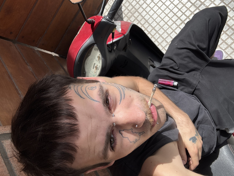 The image captures a close-up, high-angle view of a young person, likely male, reclining outdoors during bright daylight. The person is positioned on what appears to be a dark-colored scooter or moped, with its red front panel and handlebars visible behind their head.
The individual has short, dark hair with a fringe and numerous facial modifications. Visible tattoos include swirling, abstract lines with a knot-like symbol on both temples/forehead areas, and a distinct outline of what resembles a geographical map or landmass on the right cheek. Multiple facial piercings are present: two small studs below the right eye, a stud or hoop in the right nostril, two studs on the upper lip, and several spiked studs around the mouth area. The person has some stubble or light beard growth and appears to have light-colored eyes with a hint of redness around them.
In their mouth, the person holds a lit cigarette or hand-rolled joint, with smoke faintly visible from its tip. A bright pink or red lighter is resting on their dark t-shirt, near their chest. The person is wearing a dark, short-sleeved t-shirt and black pants. On their left hand, which is resting beside them, there's a visible tattoo of what appears to be a floral or intricate symbol. Part of a purple shoe or sneaker is visible under their leg.
The background shows a light-colored, square-patterned tiled ground surface to the right and a reddish-brown wooden wall or paneling to the left, indicating an outdoor setting. The bright sunlight creates strong shadows, suggesting midday. The overall impression is one of relaxation. There is no visible text in the image.