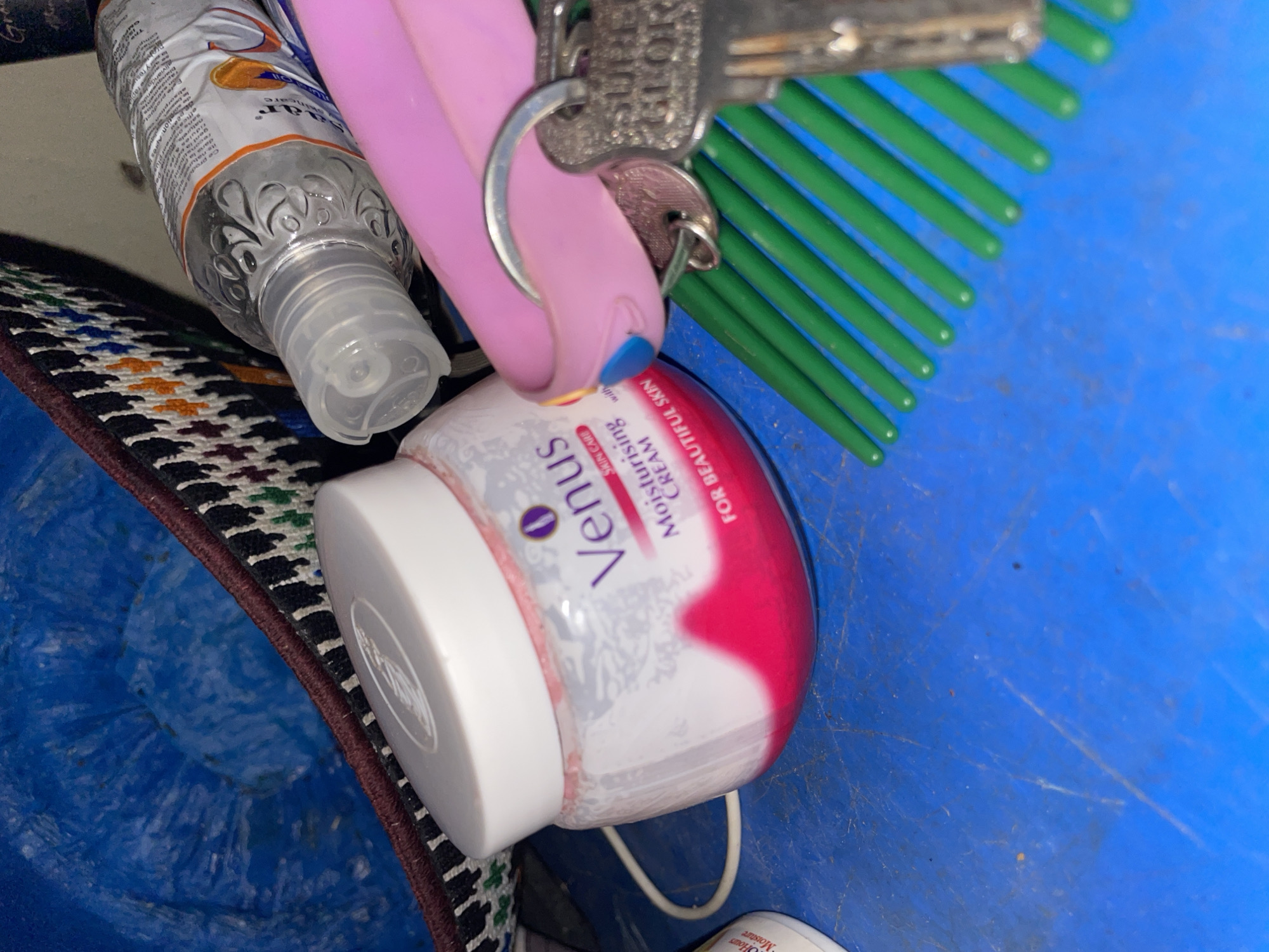 In an urban setting, possibly within Safari, Nigeria, a close-up shot captures a collection of personal care items and accessories. A jar of "Venus Moisturising Cream for Beautiful Skin" is prominently displayed, its pink and white packaging suggesting a cosmetic product. Adjacent to it, a clear plastic bottle of hand sanitizer with a light-colored cap is visible, its label partially obscured. A bright pink key holder with a light blue accent is looped around a set of keys, one of which appears to be a security key with the inscription "SURF" and "GLOR". A green plastic comb is positioned beneath these items, its teeth fanned out across a blue, textured surface that may be a tabletop or other flat surface. In the background, the edge of a woven basket with a dark trim and a pattern of multicolored threads is visible, hinting at the overall environment where these items are situated. The lighting suggests an indoor or shaded outdoor location.