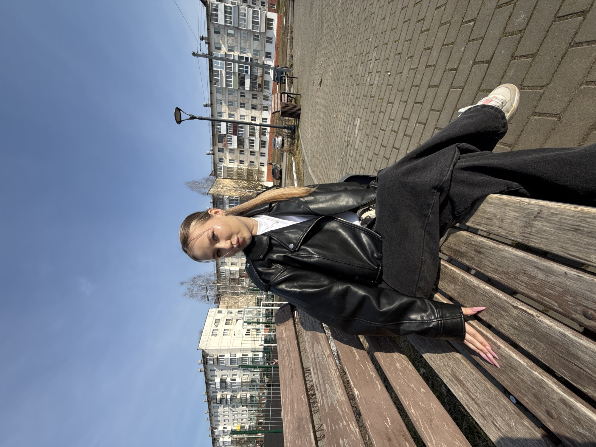 A young woman sits on a wooden park bench in Chaikovsky, Russia. She wears a black leather jacket over a white shirt and black jeans, and white sneakers. Her hair is pulled back. The background shows a blue sky, a light post, brick paving, and multi-story apartment buildings typical of Russian urban architecture.