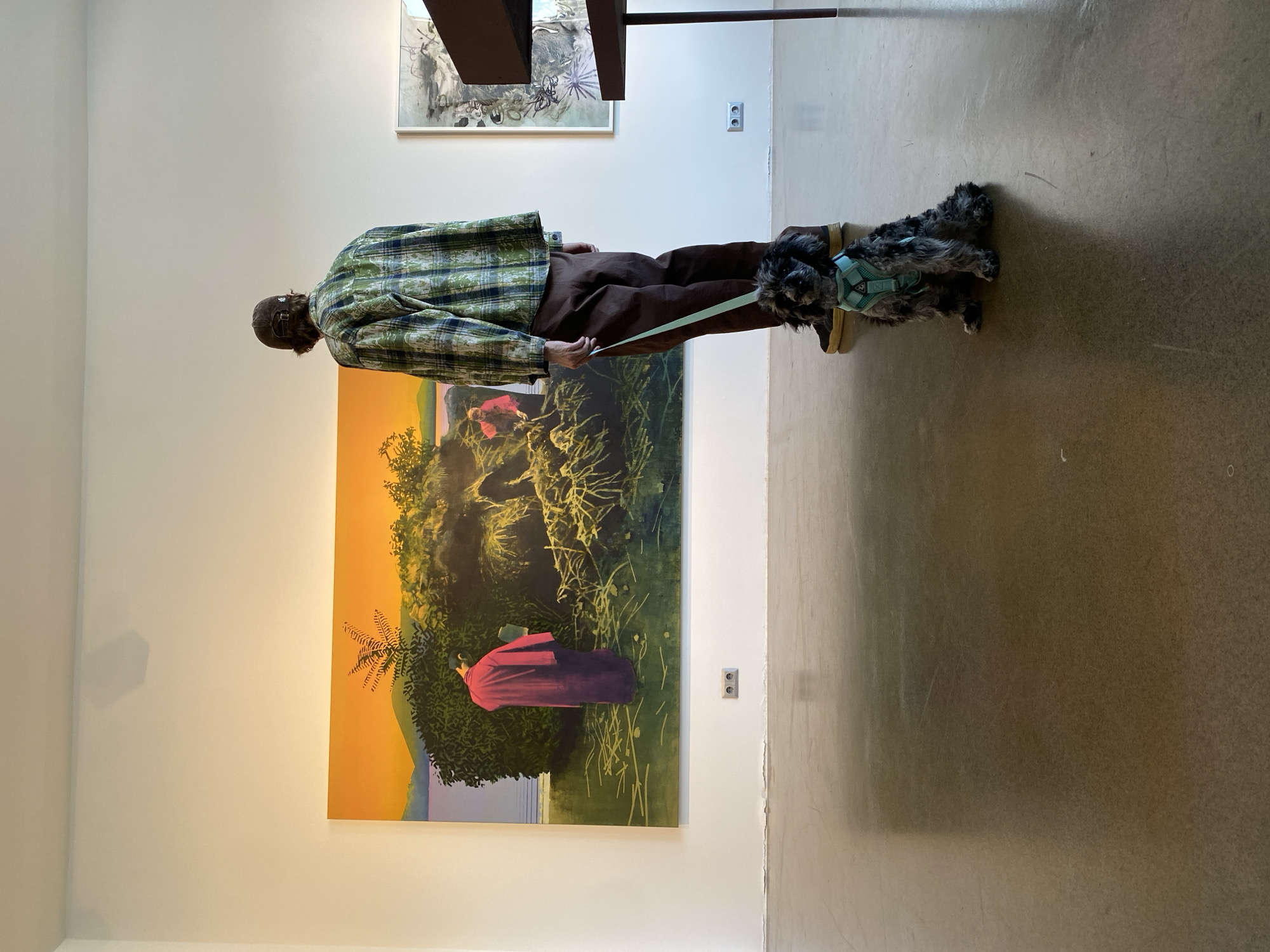 In an indoor art gallery, a person wearing a plaid shirt and brown pants stands holding a leash attached to a small, dark, curly-haired dog. The dog is seated on the polished concrete floor, facing forward. Behind the person and dog is a large, vibrant painting depicting a landscape with figures. The painting features a body of water, lush green foliage, and figures in traditional clothing. The gallery walls are white, and a shadow is cast on the wall above the painting. Another artwork, partially visible, hangs to the right. The location is identified as Saint Petersburg, Russia.