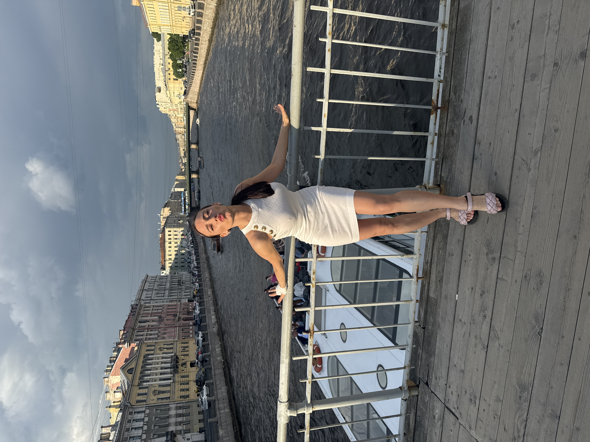 An image, rotated 90 degrees clockwise, captures a woman posing on a wooden pier or embankment overlooking a river or canal in Saint Petersburg, Russia. The woman, positioned centrally, wears a white, textured sleeveless dress with metallic button details on the straps, and light pink braided sandals. She leans with her right forearm on a white metal railing, her body slightly angled, and her left arm raised gently. Her dark hair is pulled back, and she looks towards the camera with a composed expression.
Below the railing, a white passenger boat, likely a tour vessel, is visible on the dark, calm water, with several people onboard. Across the water, a cityscape unfolds, featuring rows of multi-story buildings with varied architectural styles and colors, lining the embankment. Cars are parked along the street. The sky above is largely overcast with grey clouds, suggesting a cloudy day, though the lighting on the woman and buildings is bright. No specific text is clearly discernible in the image.